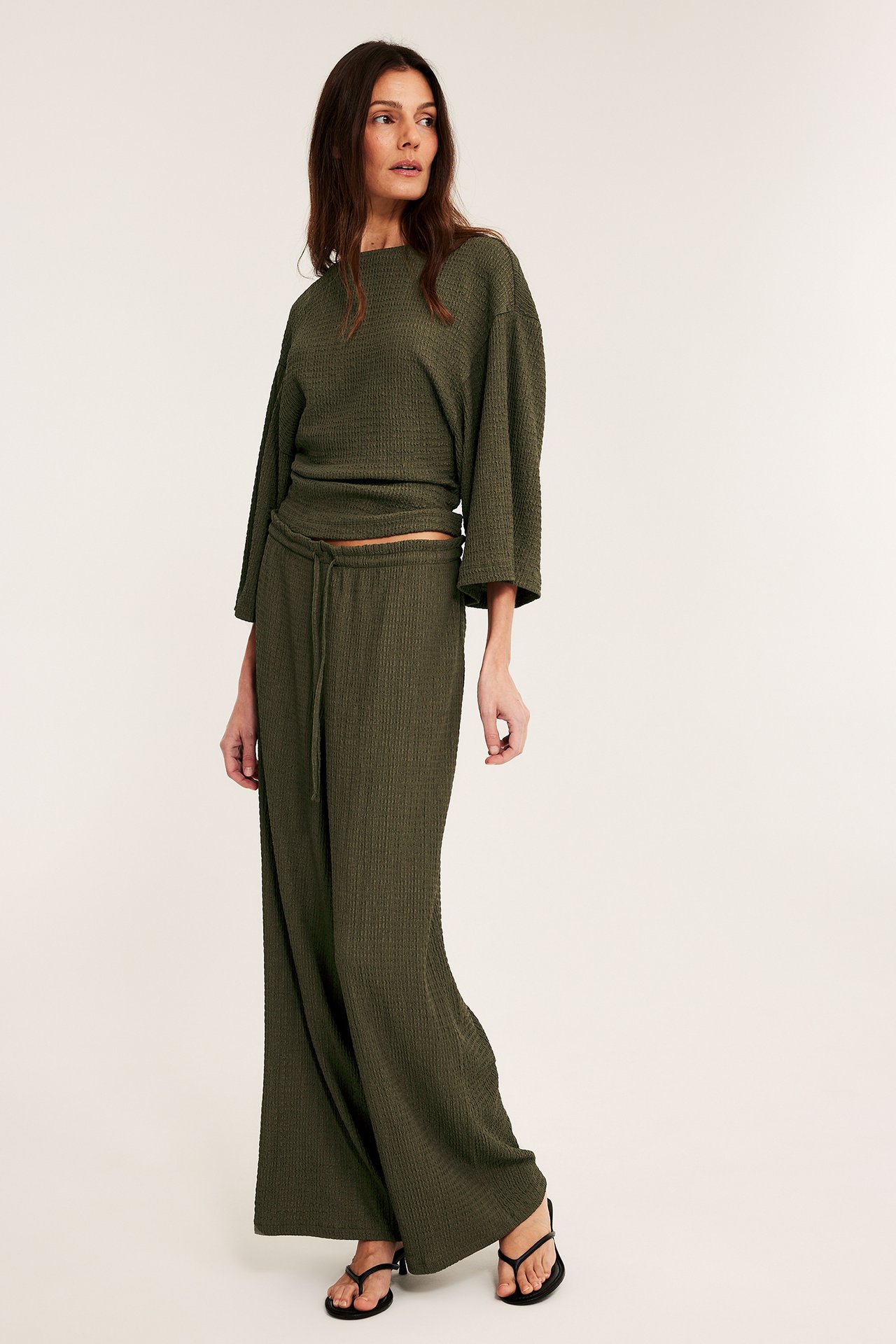 Framifrån: Kvinna i gröna dragskobyxor i strukturerad jersey, matchande topp, svarta sandaler.