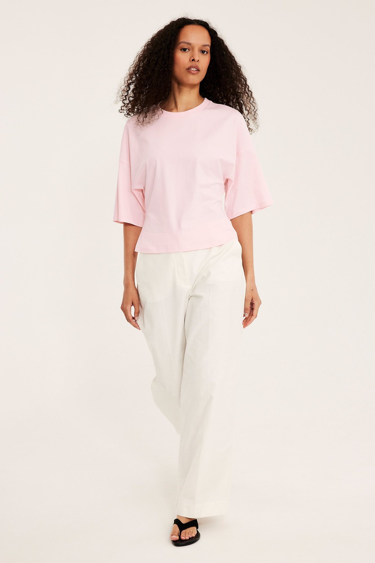 Framifrån: kvinna i ljusrosa t-shirt med droppade axlar, ecruvida byxor och svarta sandaler.
