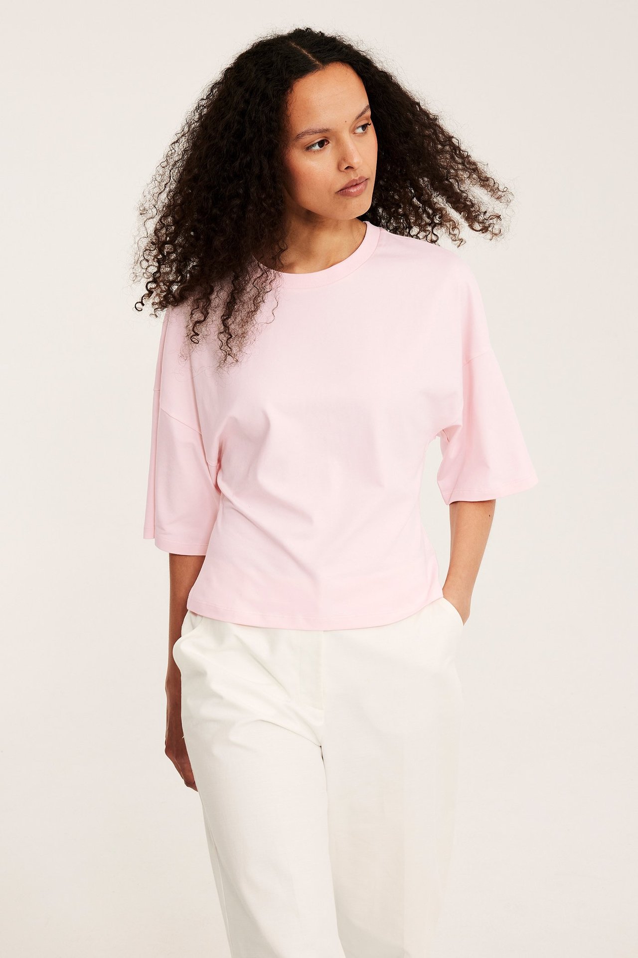 Framifrån: kvinna i ljusrosa figurnära t-shirt i bomull med nedhasade axlar och vita byxor.
