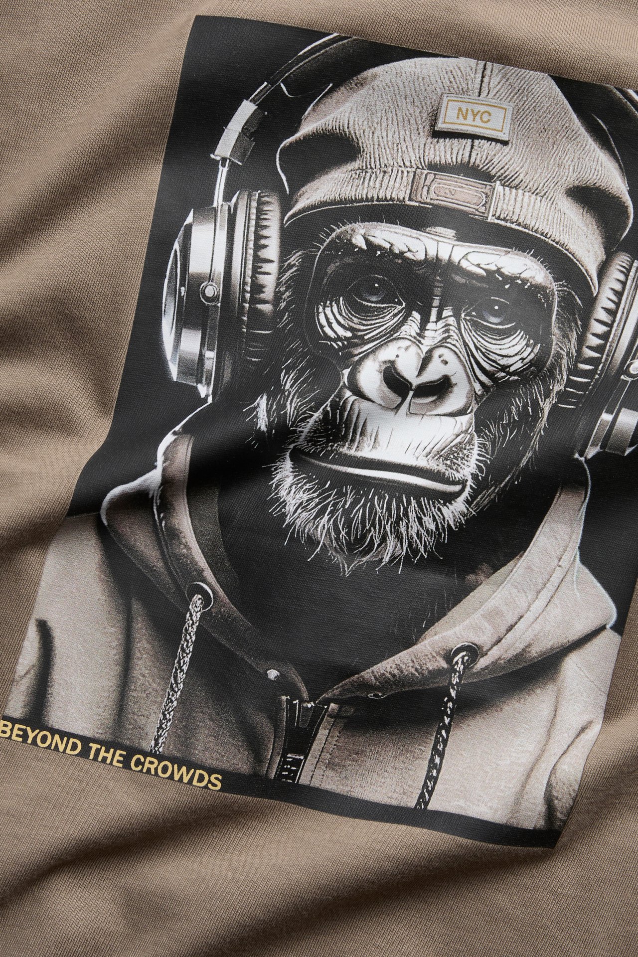 Zbliżenie brązowego t-shirtu z grafiką szympansa w czapce, słuchawkach i napisem 'BEYOND THE CROWDS'.