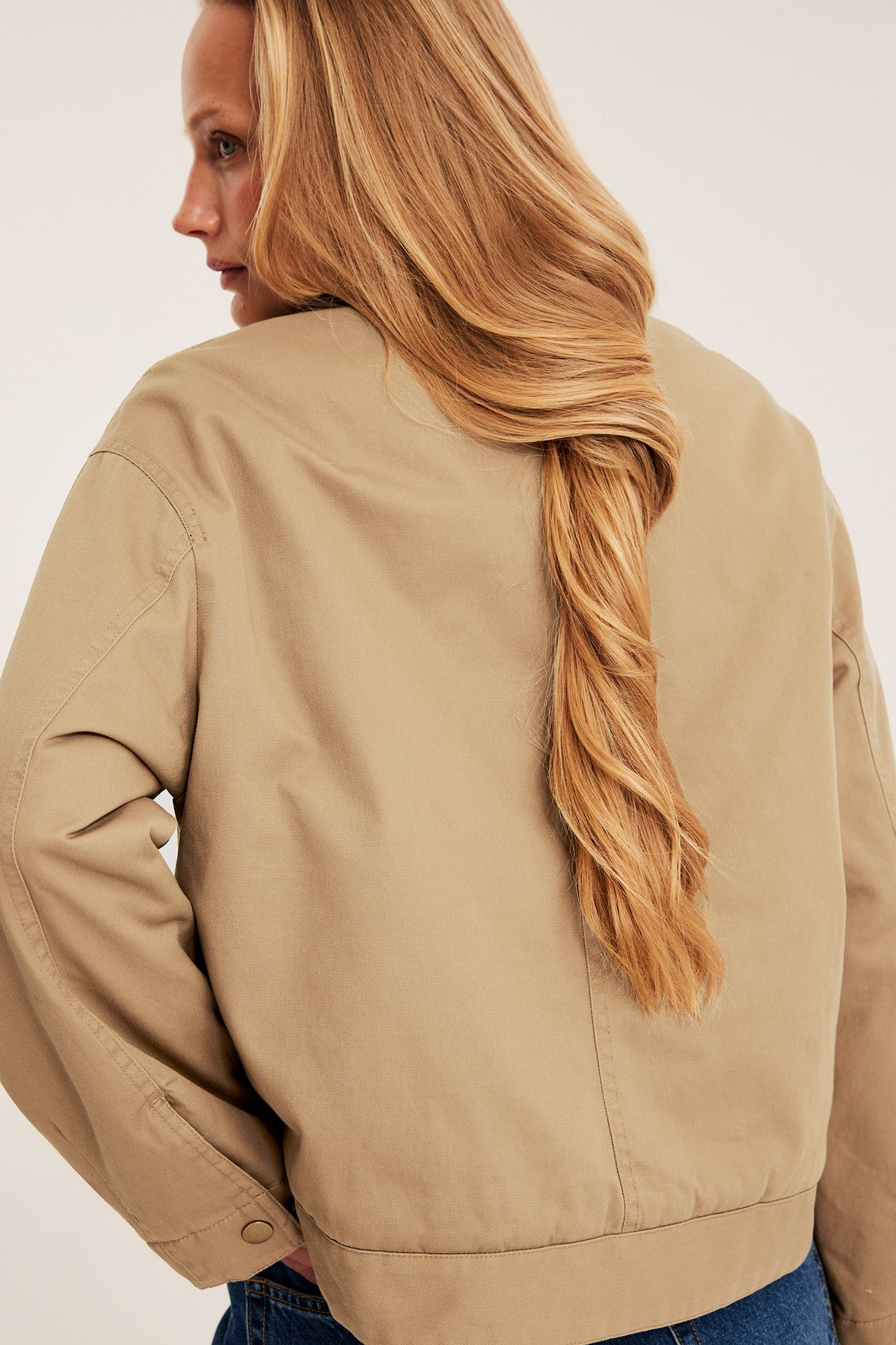 Rygg-sidovy av en kvinna i en kort beige canvasjacka med knäppta ärmslut och blå jeans.