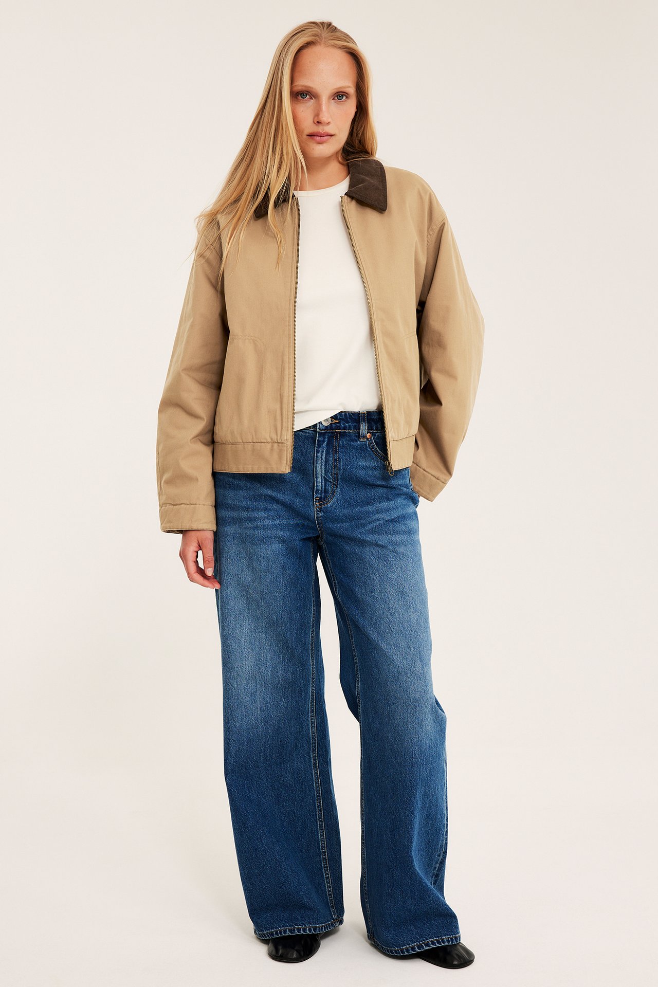 Kvinna bär beige canvasjacka med brun manchesterkrage, vit t-shirt och blåa vida jeans.