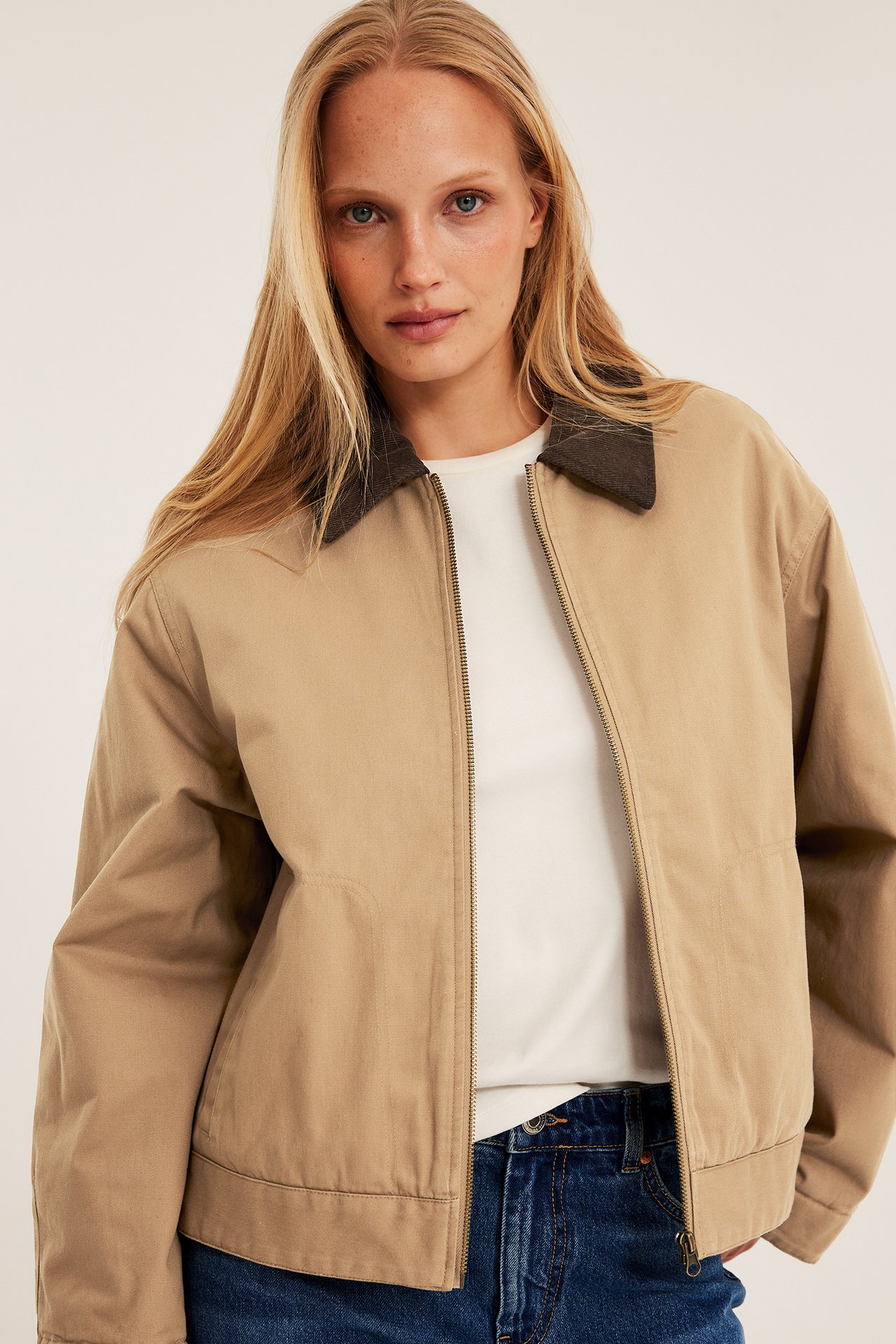 Framsida: Kvinna bär beige kort canvasjacka med mörkbrun manchesterkrage, vit t-shirt, blåa jeans.