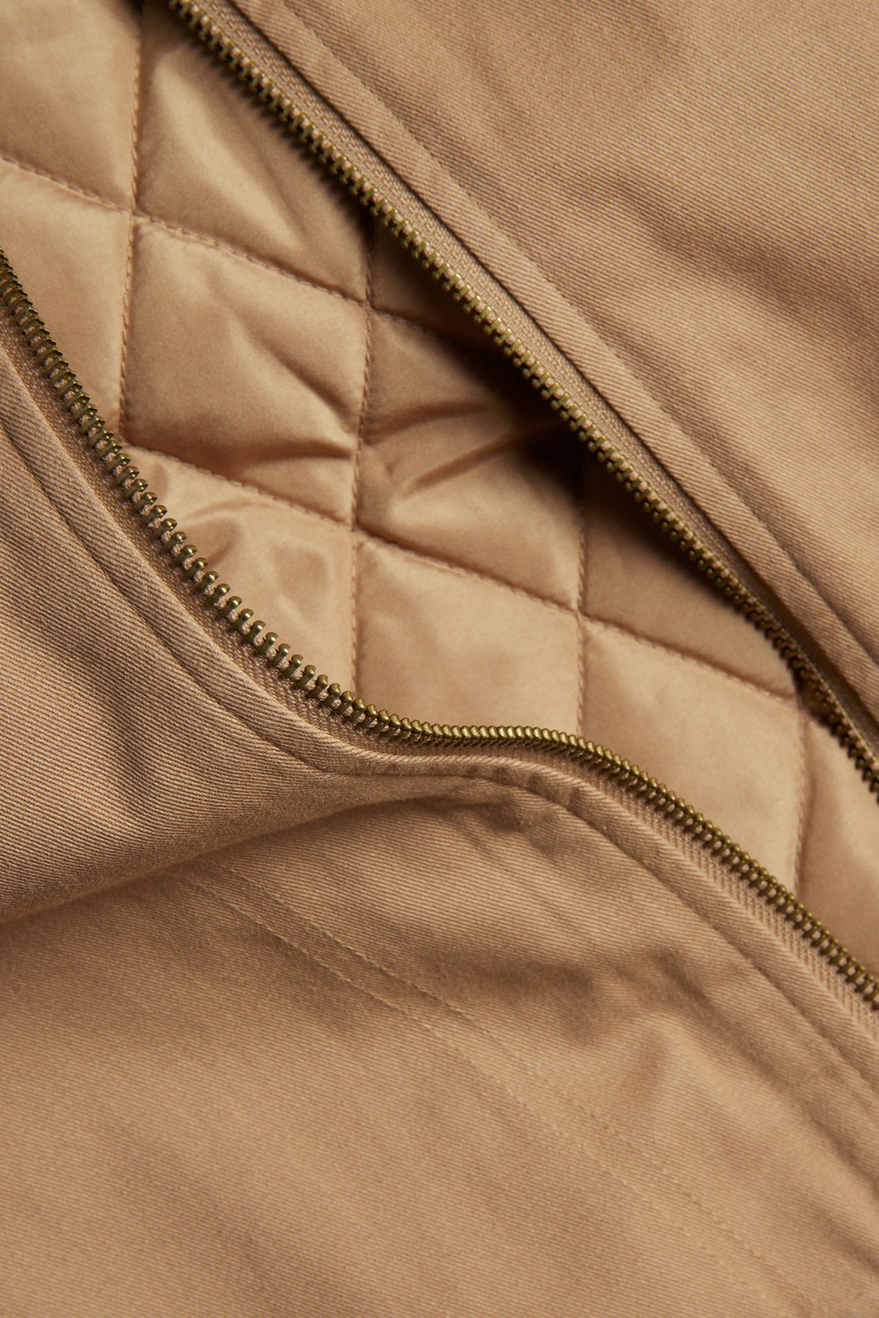 Närbild av en beige canvasjacka med bronsfärgad dragkedja och quiltat beigt foder.