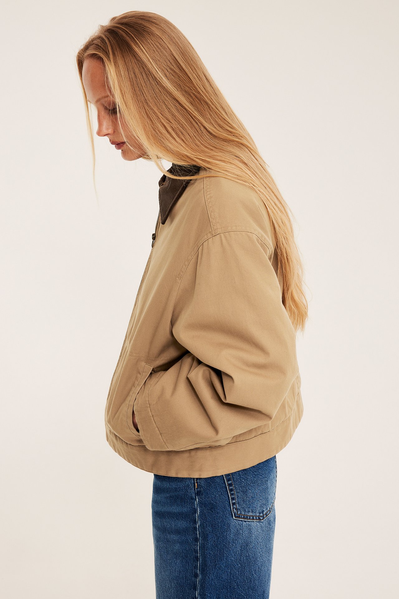 Sidovy av en kvinna i beige kort canvasjacka med brun manchesterkrage och blå jeans.
