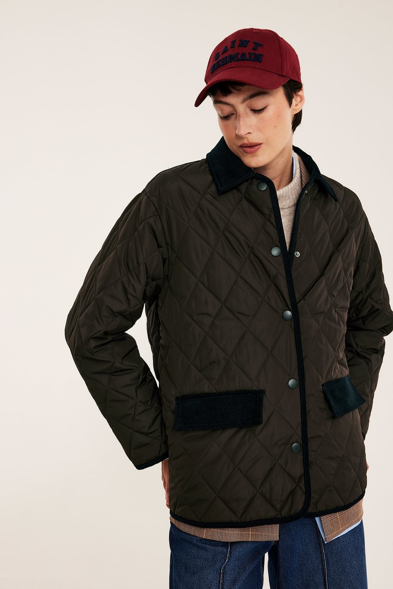 Kvinnelig modell bærer en mørk oliven quiltet jakke, vinrød caps, beige genser og blå jeans.