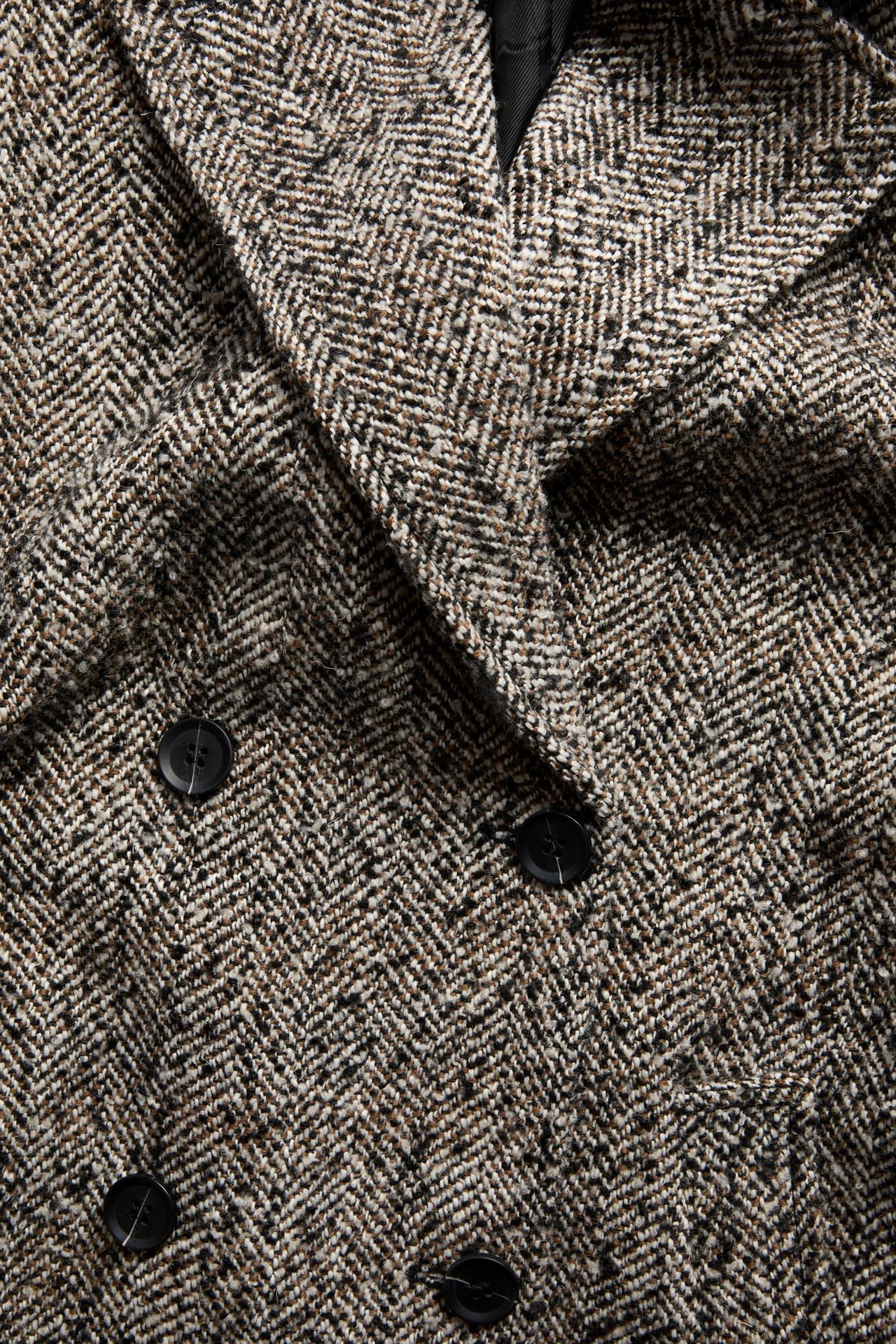 Närbild på en svart, vit och brun dubbelknäppt kappa i fiskbensmönstrad tweed med svarta knappar.