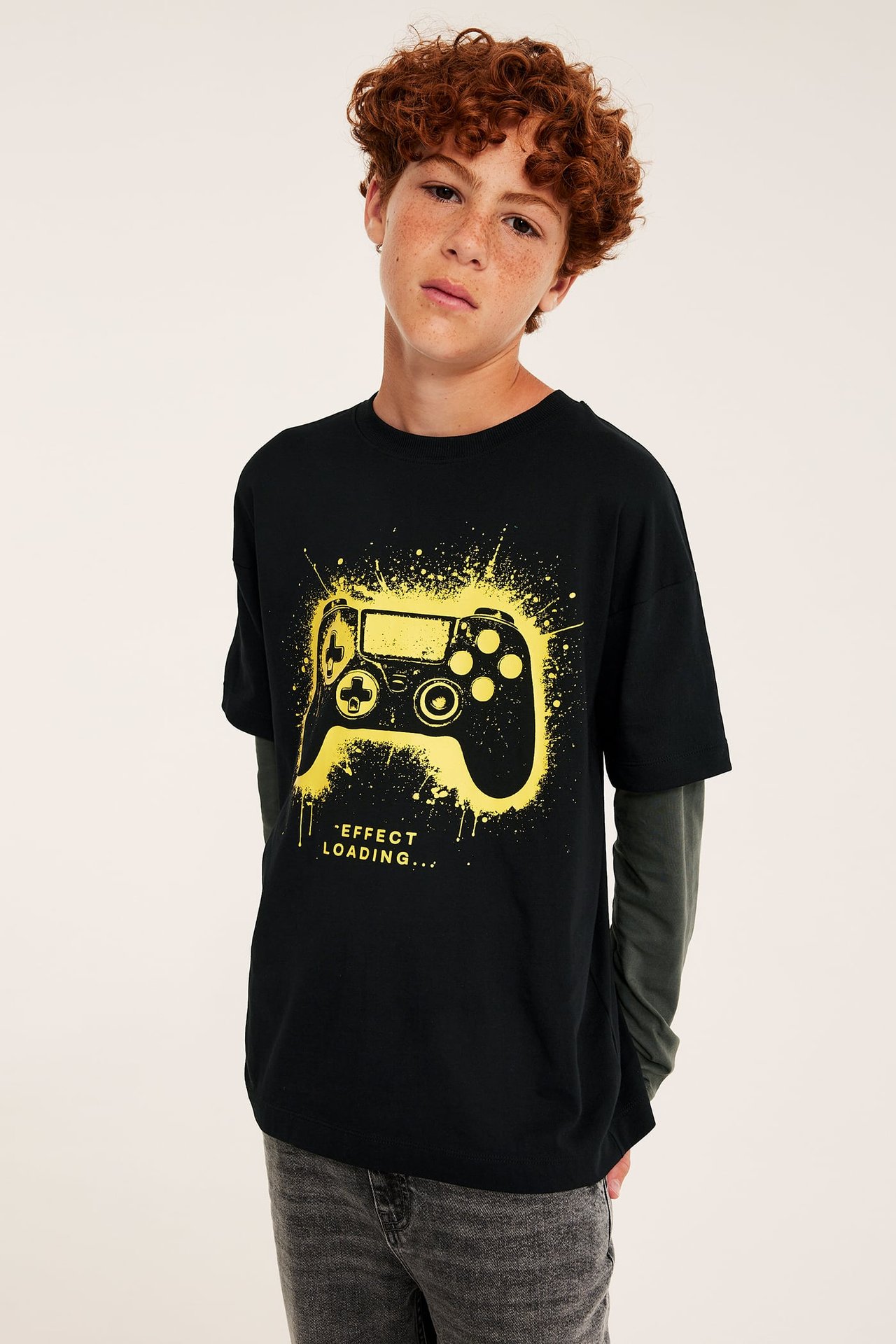 Dziecko w czarnym T-shircie z żółtym nadrukiem gamepada, na zielonej bluzce i szarych jeansach.
