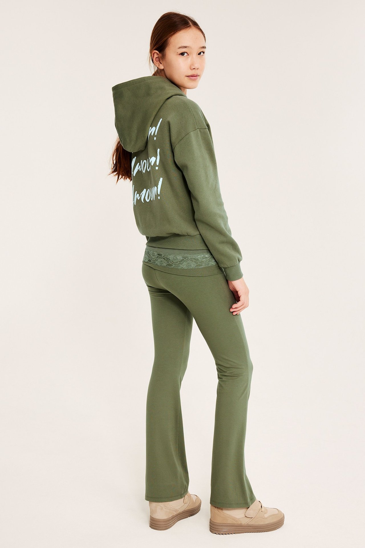 Widok z boku: dziecko w ciemnozielonych rozszerzanych legginsach z koronką, pasującej bluzie i beżowych butach.