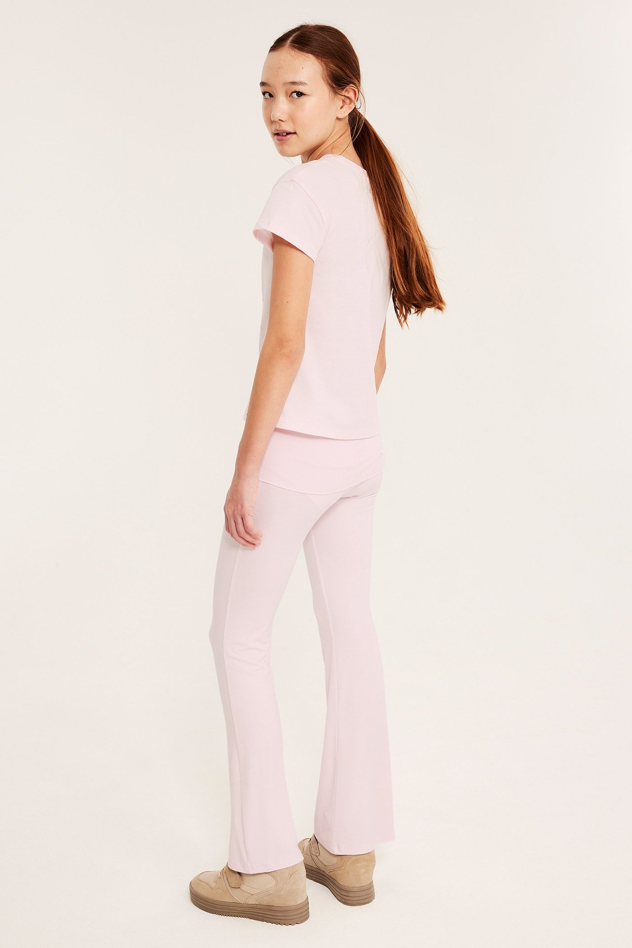 Widok z boku, od tyłu: dziecko w jasnoróżowych rozszerzanych spodniach do jogi, pasującym t-shircie i beżowych sneakersach.