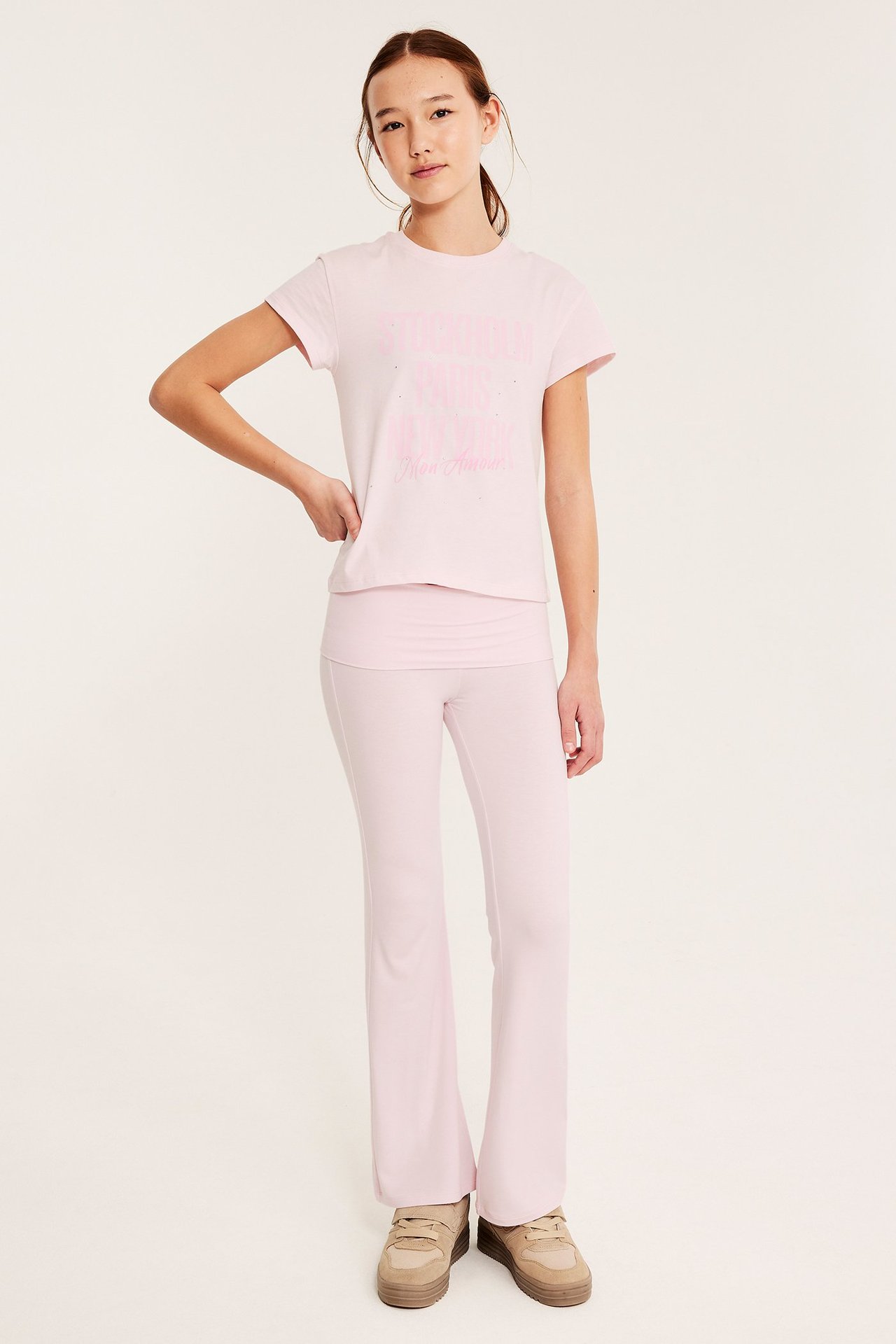 Edestä: Lapsi vaaleanpunaisissa levenevissä joogahousuissa taitettavalla vyötäröllä, pinkki t-paita ja beiget lenkkarit.