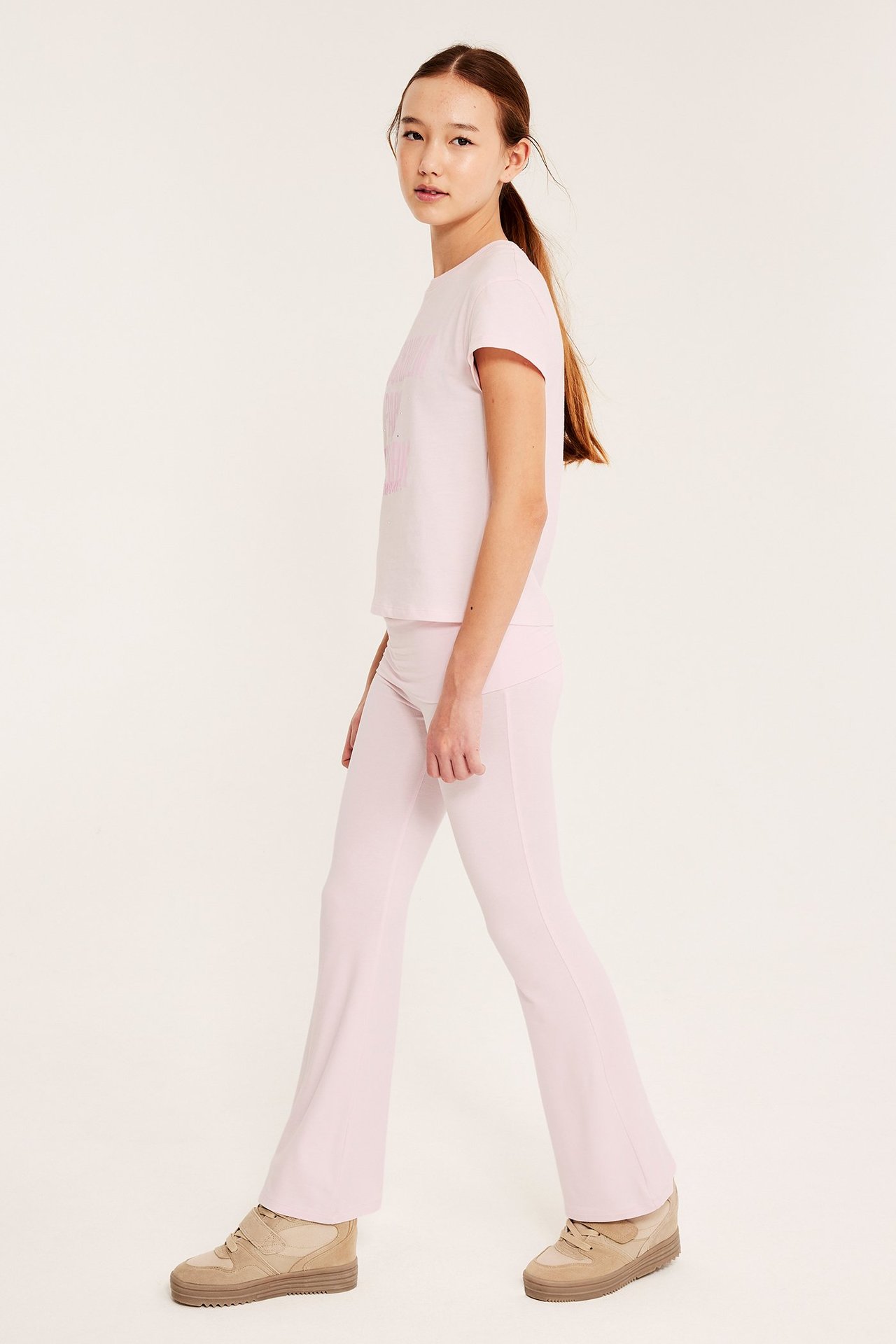 Widok z boku dziecka w jasnoróżowych legginsach, jasnoróżowej koszulce i beżowych trampkach.