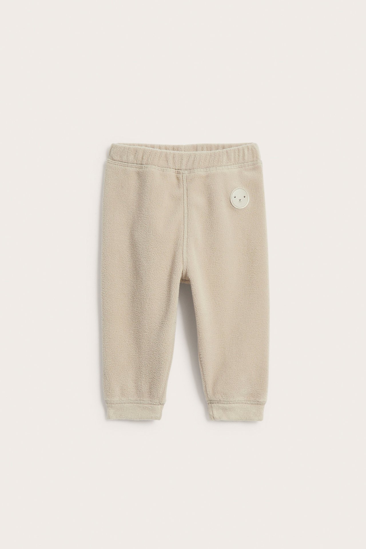 Frontvy av beige babyfleecebyxor med resår i midjan och ett litet vitt märke.