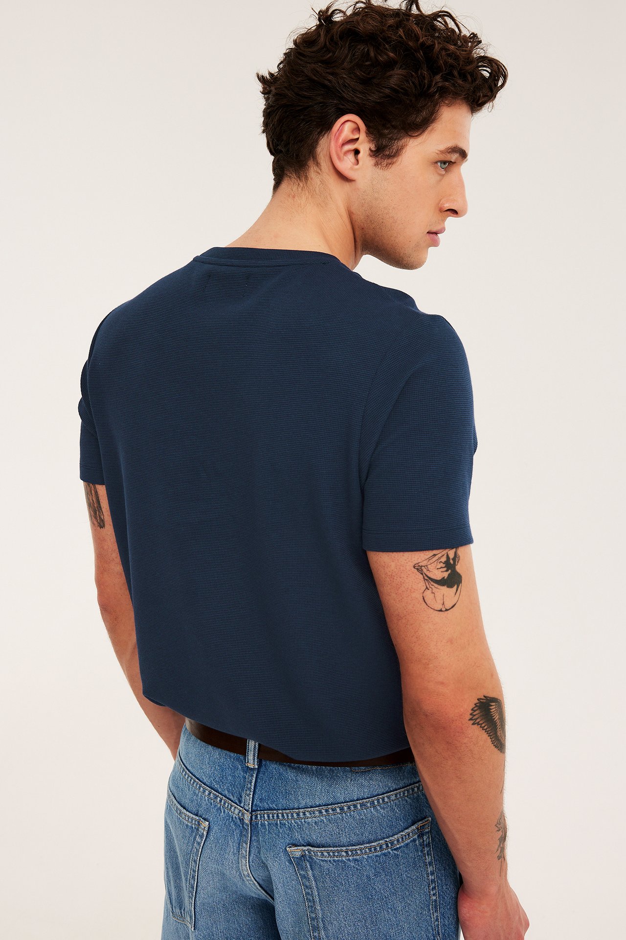 Widok z tyłu na mężczyznę w granatowej, strukturalnej koszulce z krótkim rękawem, jasnych jeansach i brązowym pasku.