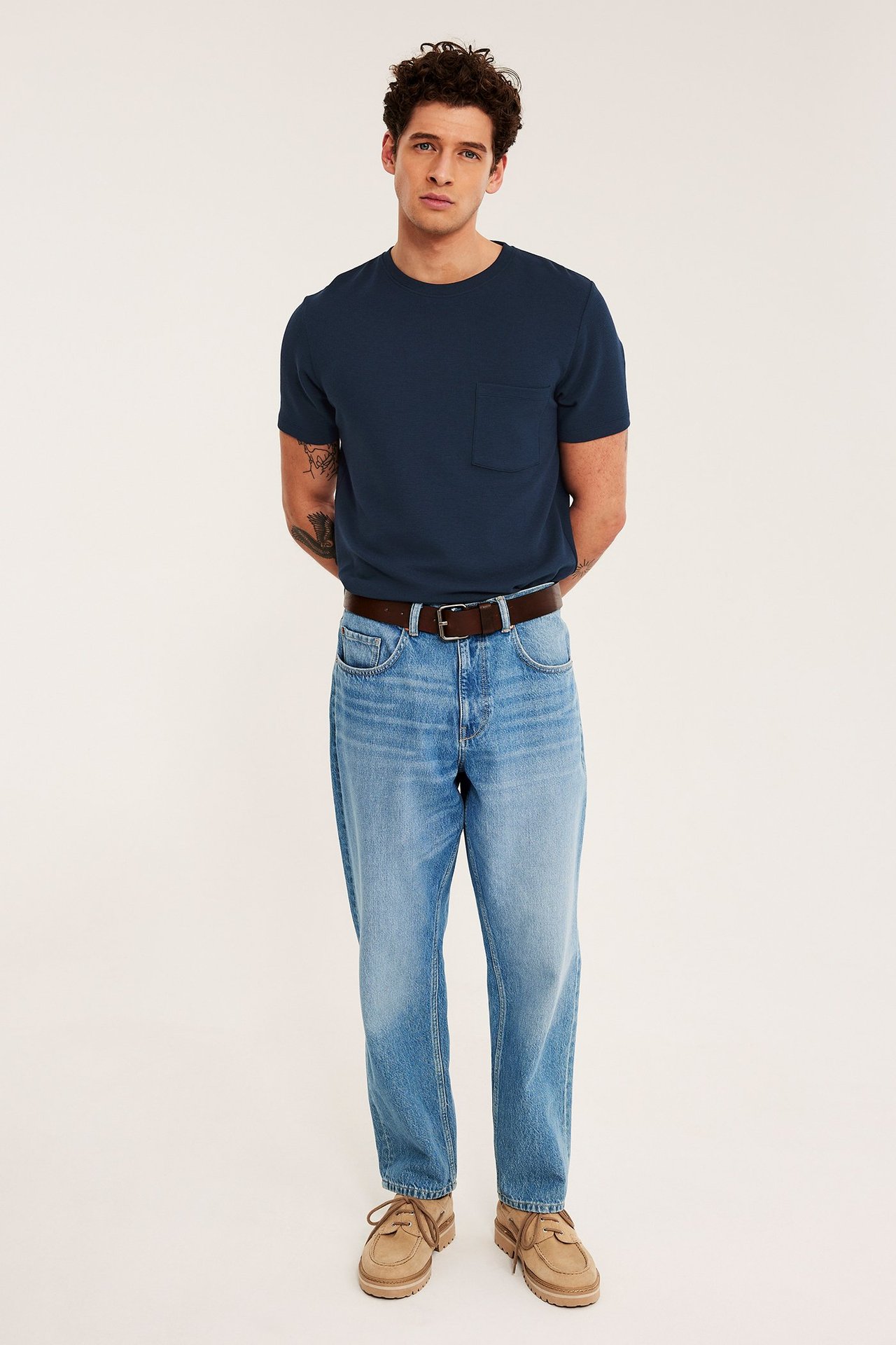 Forfra mann i mørkeblå strukturert T-skjorte med brystlomme, lyseblå jeans, brunt belte og beige sko.