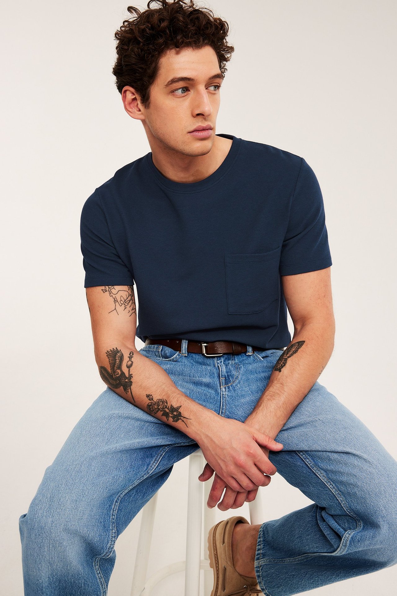 Mann i mørkeblå strukturert t-skjorte med brystlomme, lyseblå jeans, brunt belte og beige loafers.