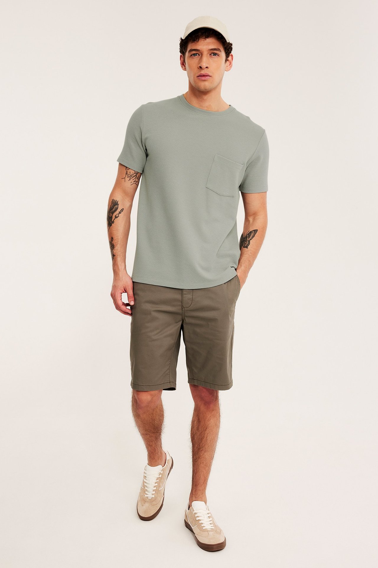Mann i lys grønn strukturert t-skjorte med brystlomme, khakifargede shorts, beige sneakers og lys beige caps.