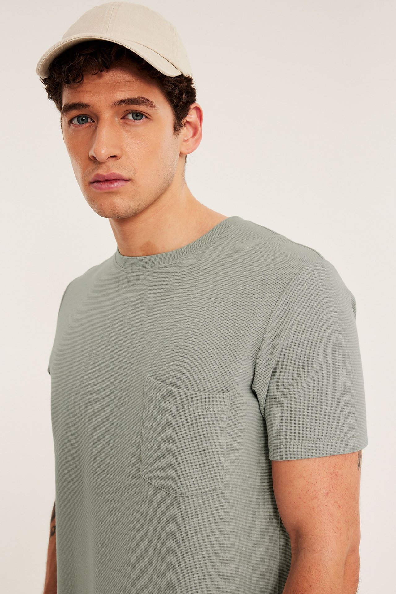 Edestä, mies yllään vaaleanvihreä kuvioitu t-paita rintataskulla ja beigenvärinen lippalakki.