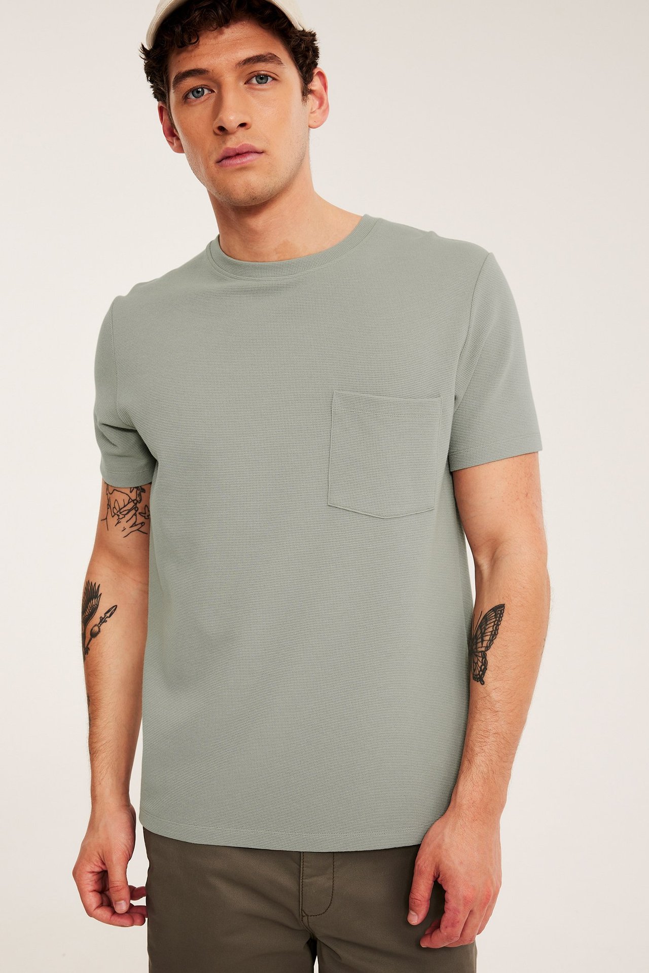 Edestä: Mies pukeutunut vaaleanvihreään kuvioituun t-paitaan rintataskulla, tummiin housuihin ja beigeen lippikseen.