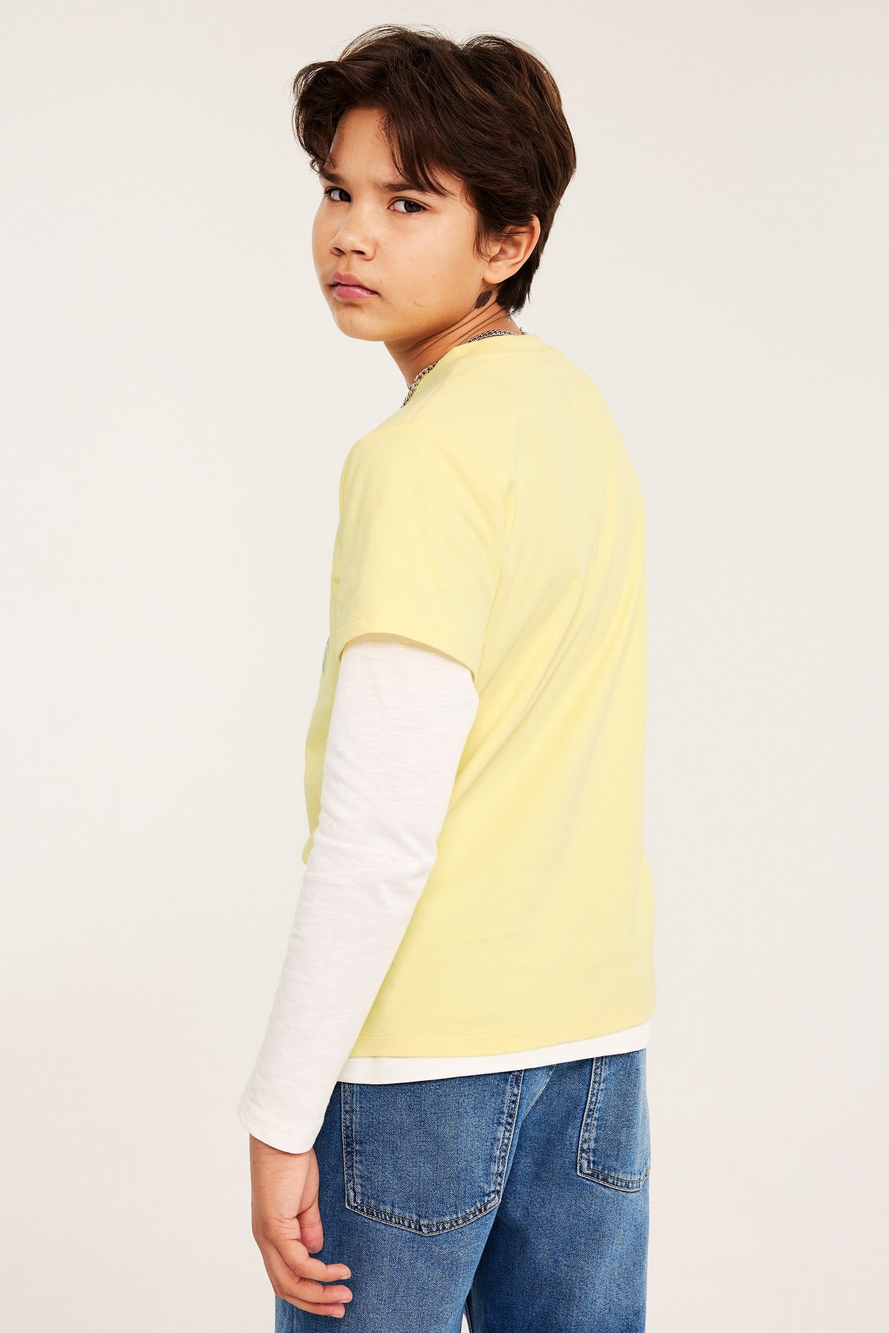 Sidebilde av et barn i en gul T-skjorte over en hvit langermet genser og blå jeans.