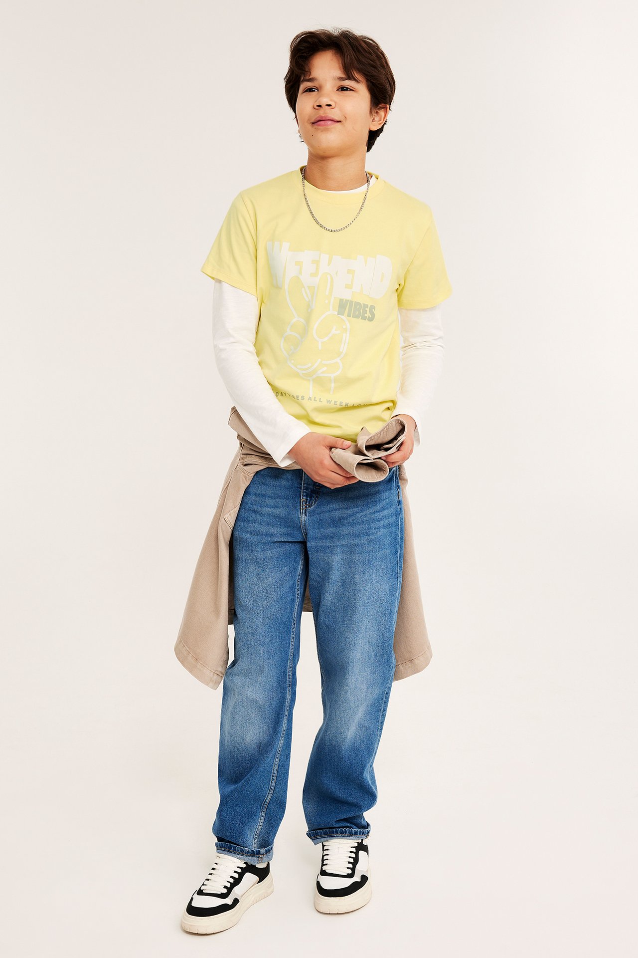 Frontvy av ett barn i gulmönstrad T-shirt över vit långärmad tröja, blå jeans och sneakers.