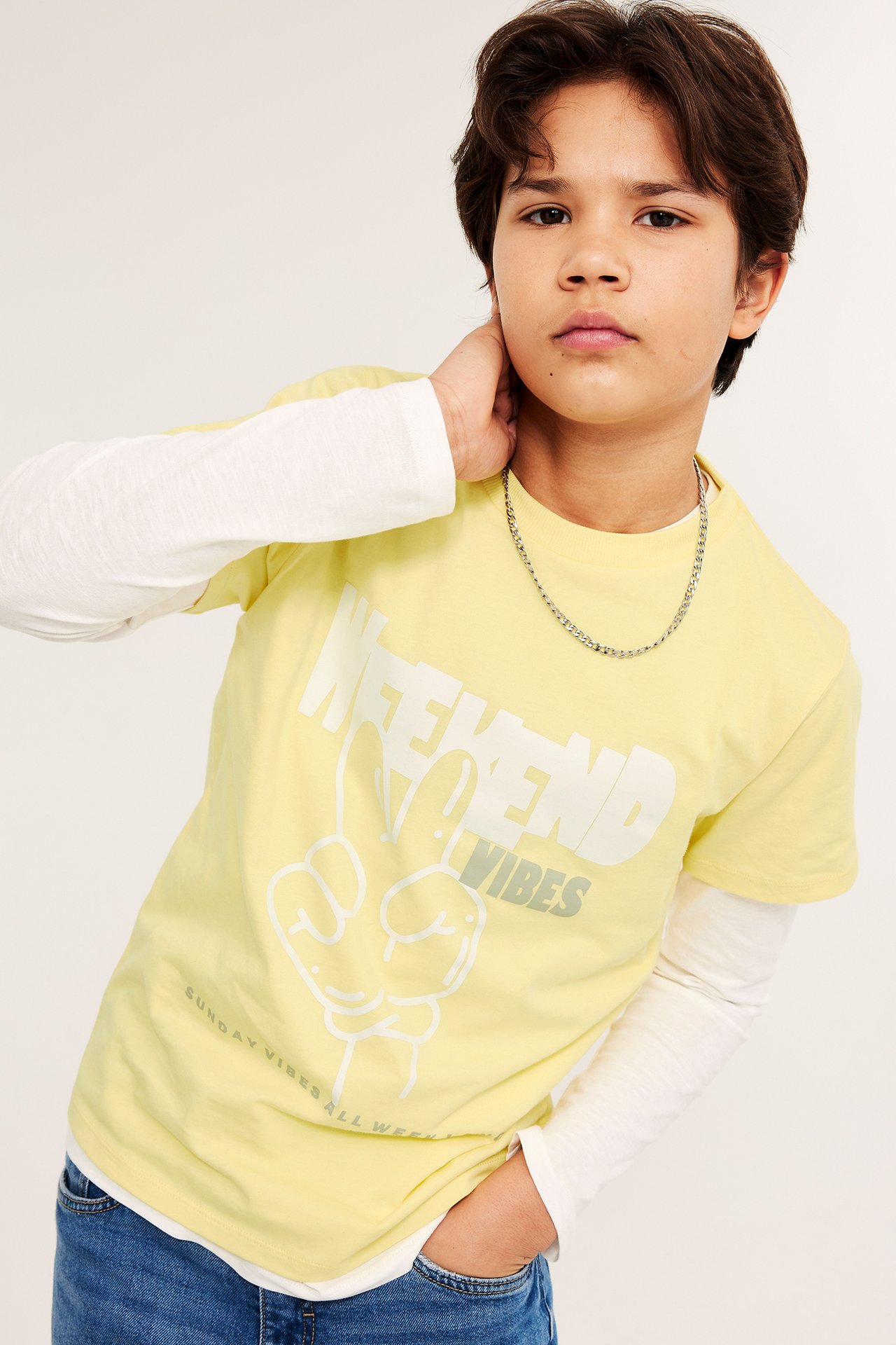 Forfra: Barn i gulmønstret T-skjorte over hvit langermet, blå jeans og et sølvkjede.