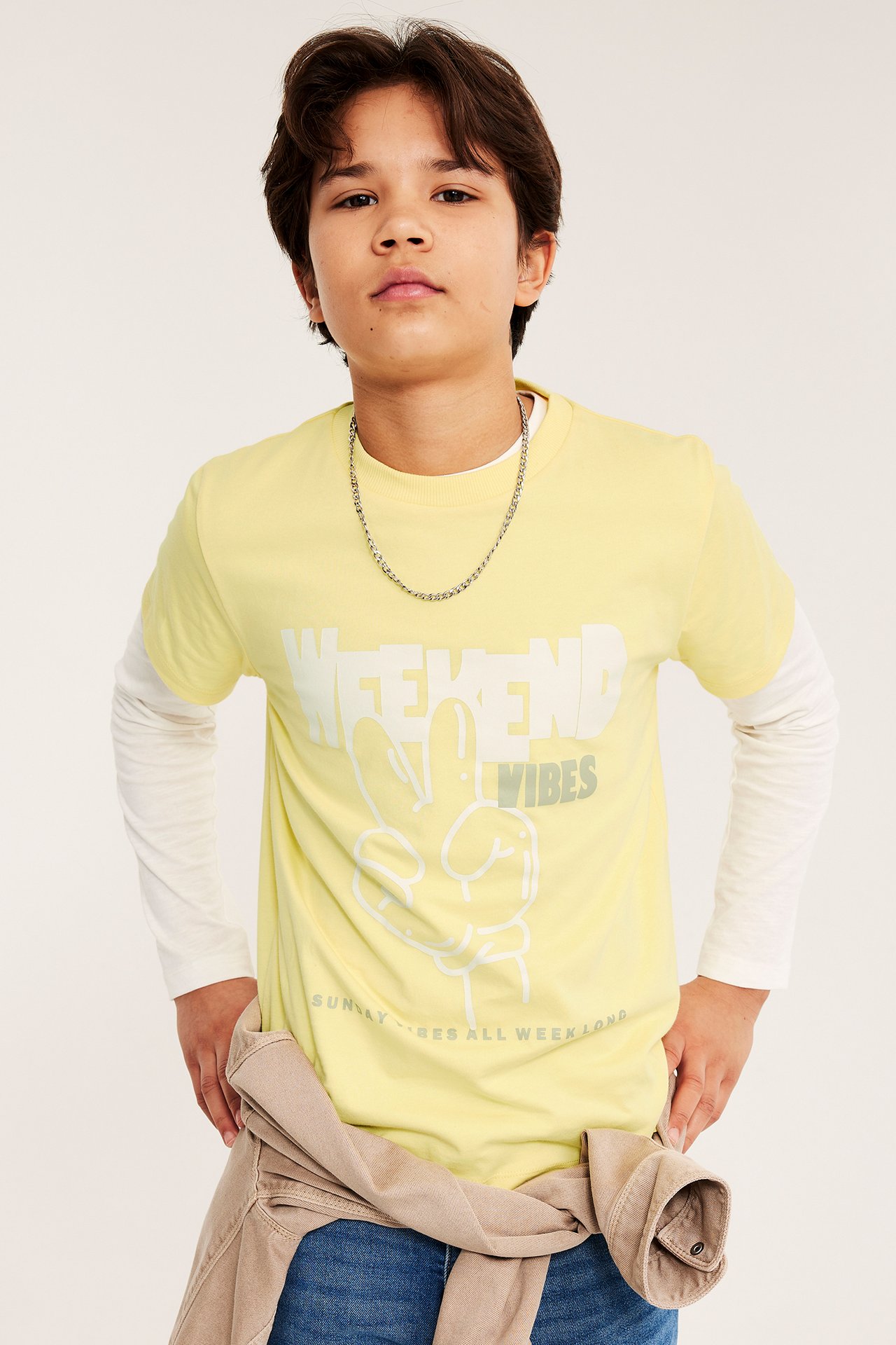Framifrån på ett barn i gul bomulls-T-shirt med 'Weekend Vibes'-tryck över vit långärmad tröja.