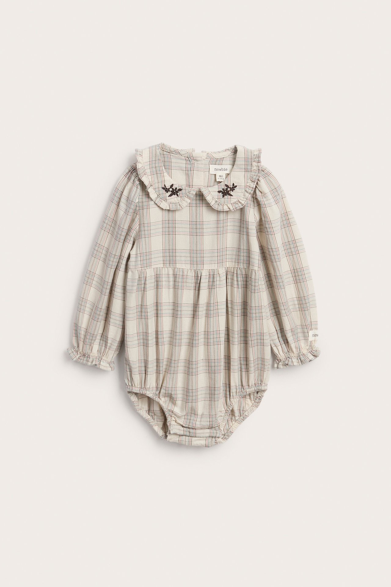 Framsida av rutig babybody i krämvitt med lång ärm och broderad volangkrage.