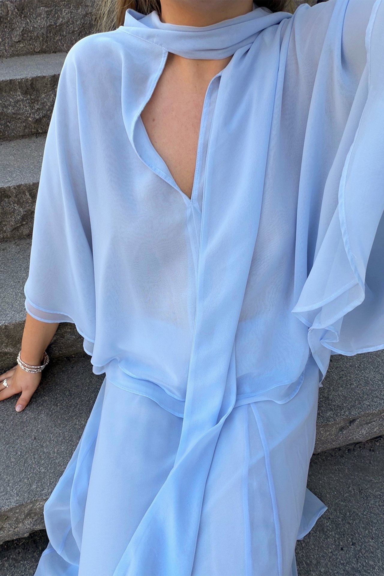 Lähikuva naisesta vaaleansinisessä sifonkikaapupuserossa, yhteensopivissa housuissa ja hopearannekoruissa.