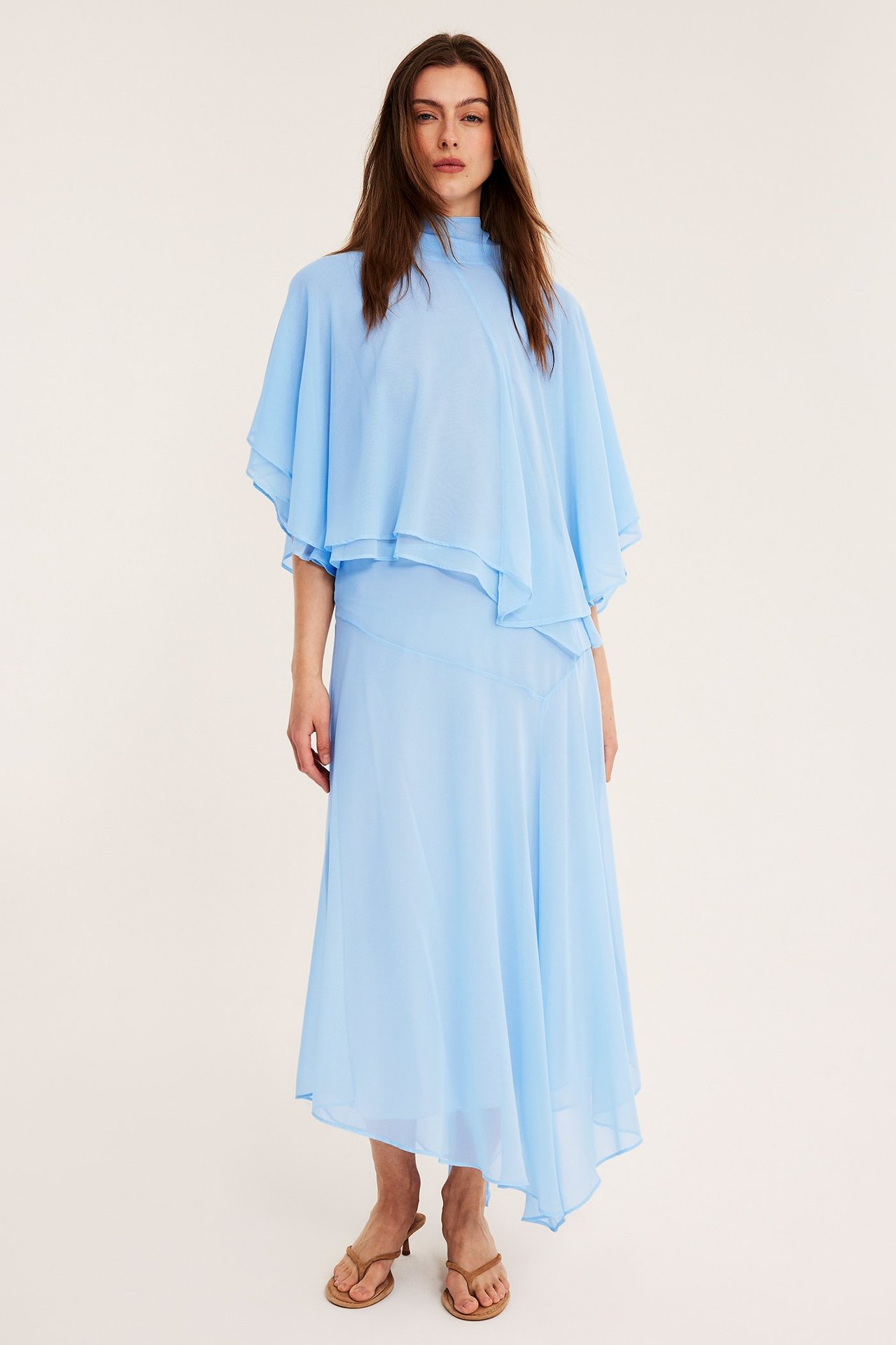 Edestäpäin, nainen yllään vaaleansininen sifonki cape-pusero, yhteensopiva epäsymmetrinen hame ja ruskeat korkeakorkoiset sandaalit.