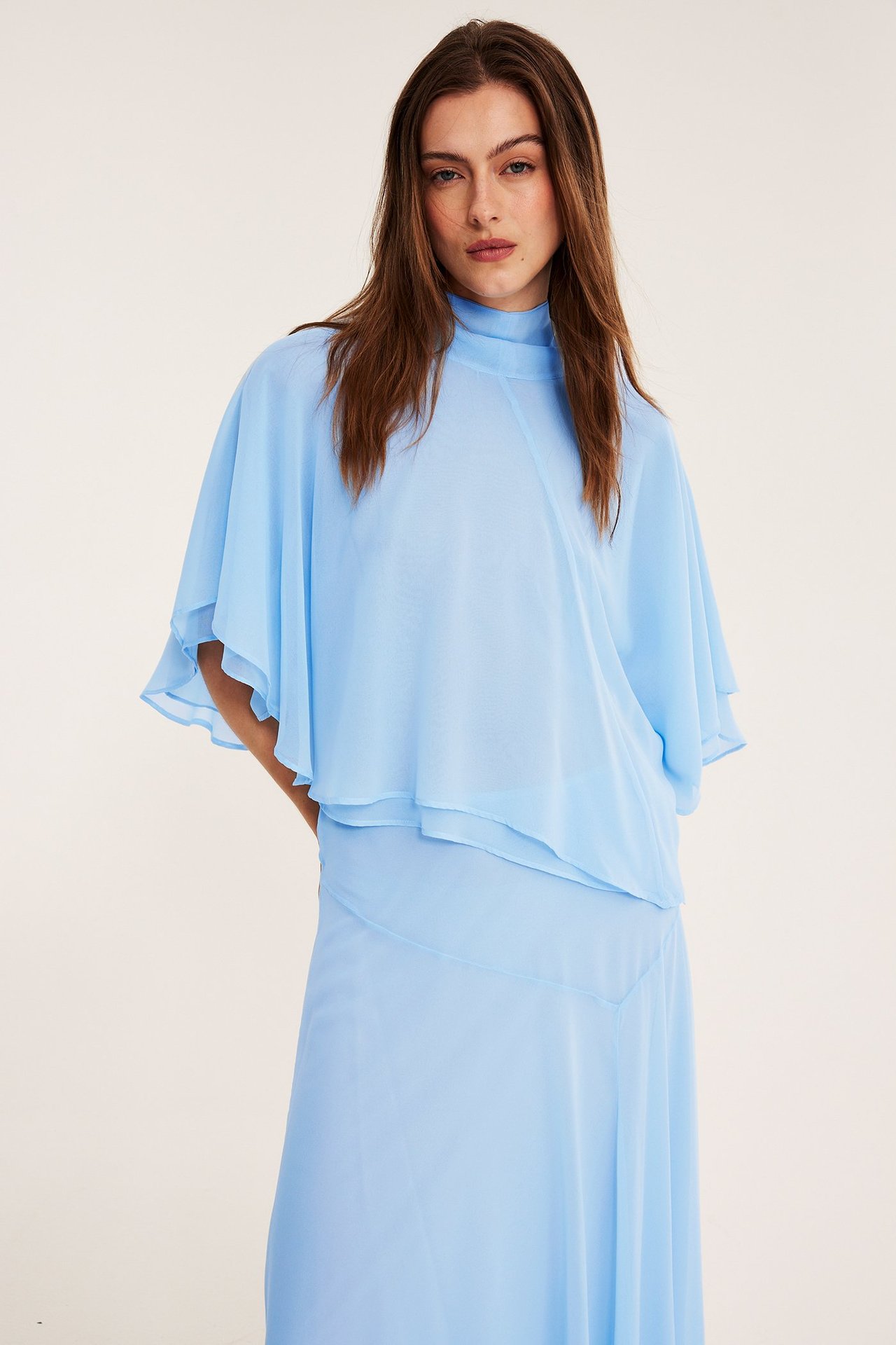 Widok z przodu, kobieta w jasnoniebieskiej szyfonowej bluzce z peleryną, stójką i pasującej spódnicy.