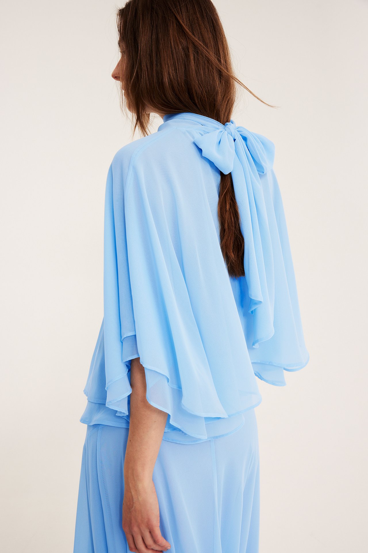 Kvinnas sidovy: ljusblå chiffongblus med cape-silhuett, rosett bak, matchande kjol.
