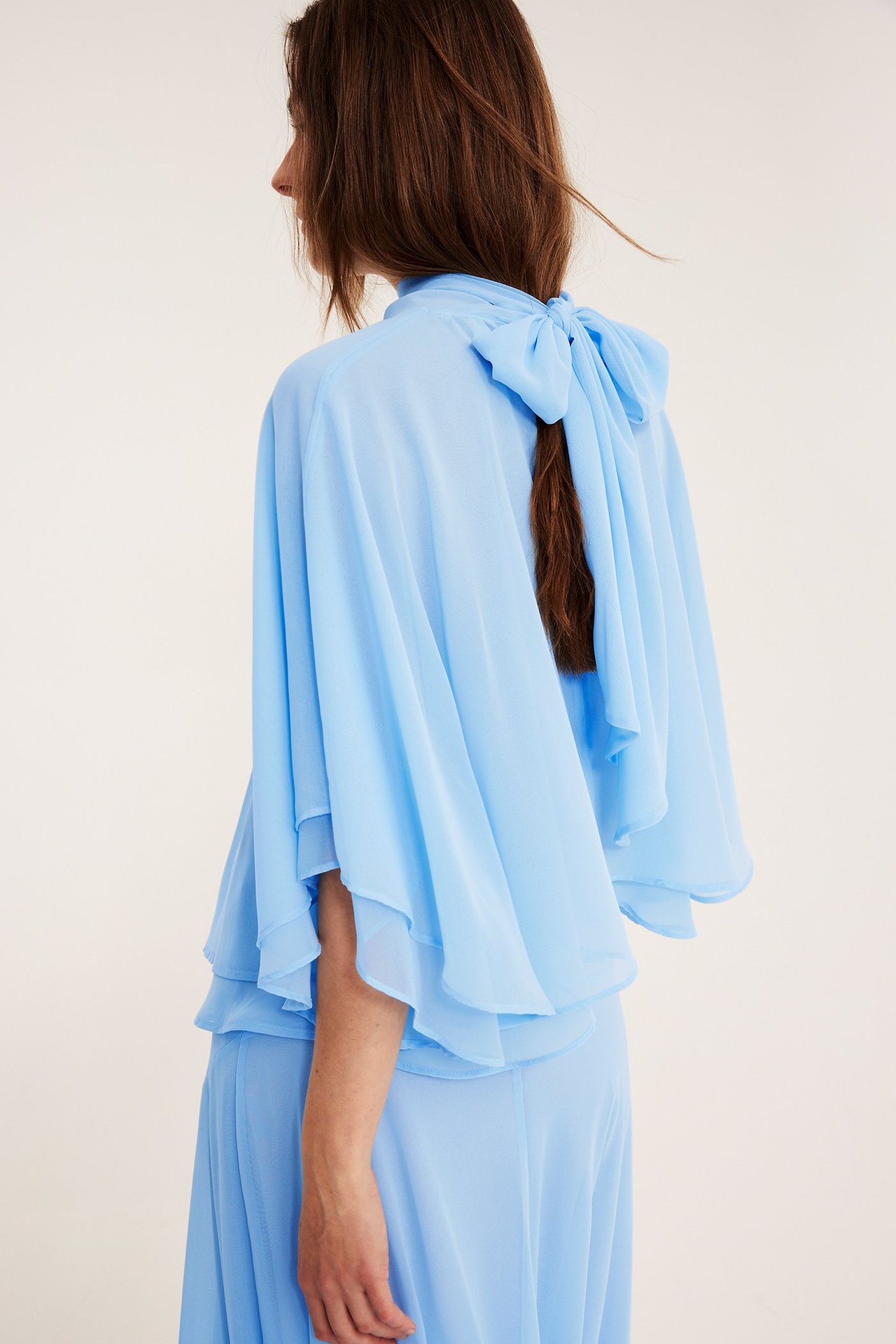 Widok z boku kobiety: jasnoniebieska szyfonowa bluzka-peleryna z kokardą z tyłu, z pasującą spódnicą.