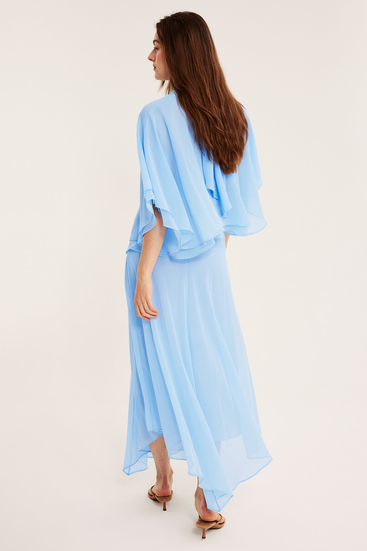 Kvinna bakifrån i en ljusblå asymmetrisk lång chiffongkjol, matchande topp och bruna sandaletter.