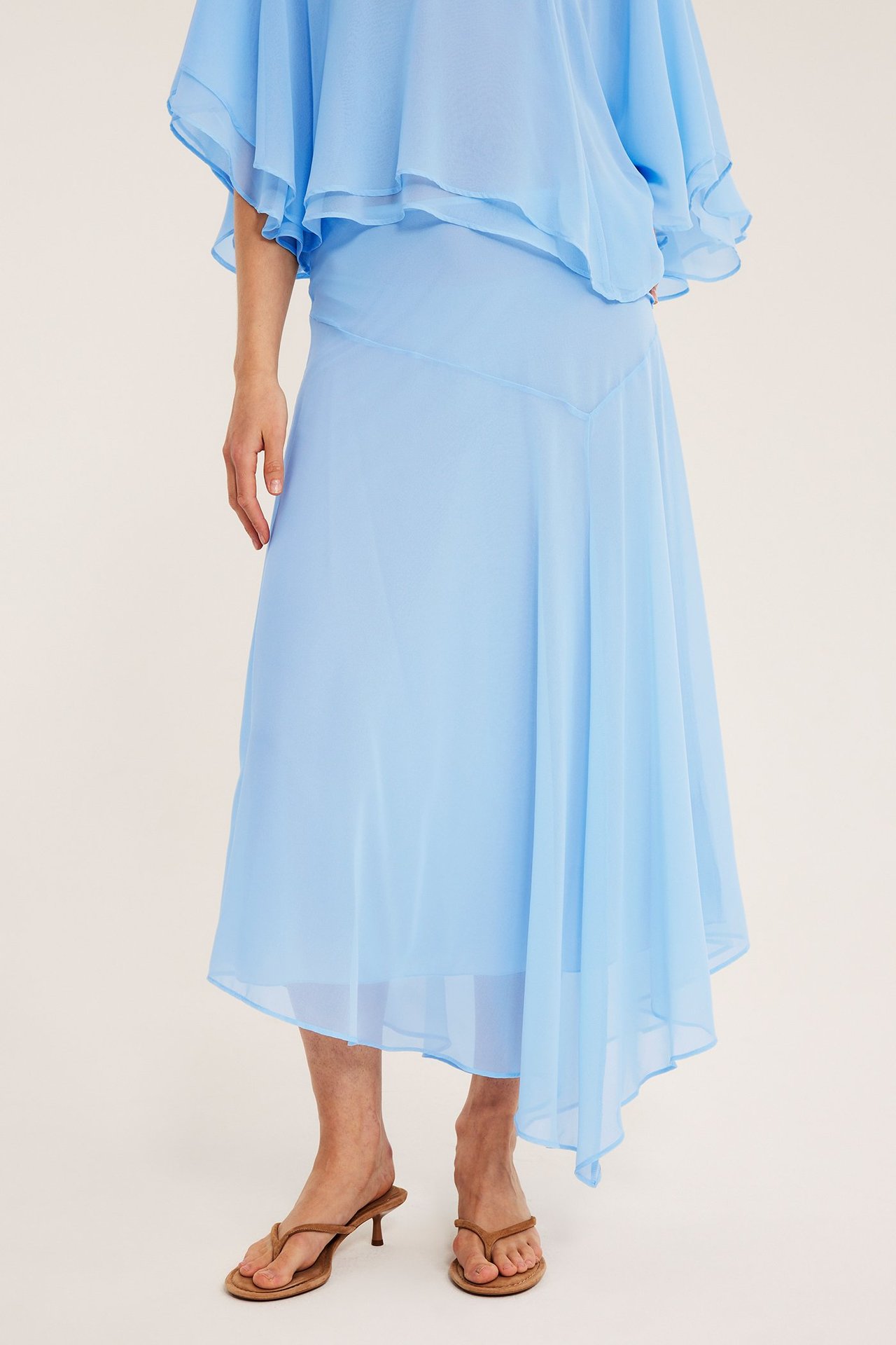Framifrån, kvinna bär ljusblå asymmetrisk chiffongkjol, matchande topp och bruna tåremssandaler med klack.