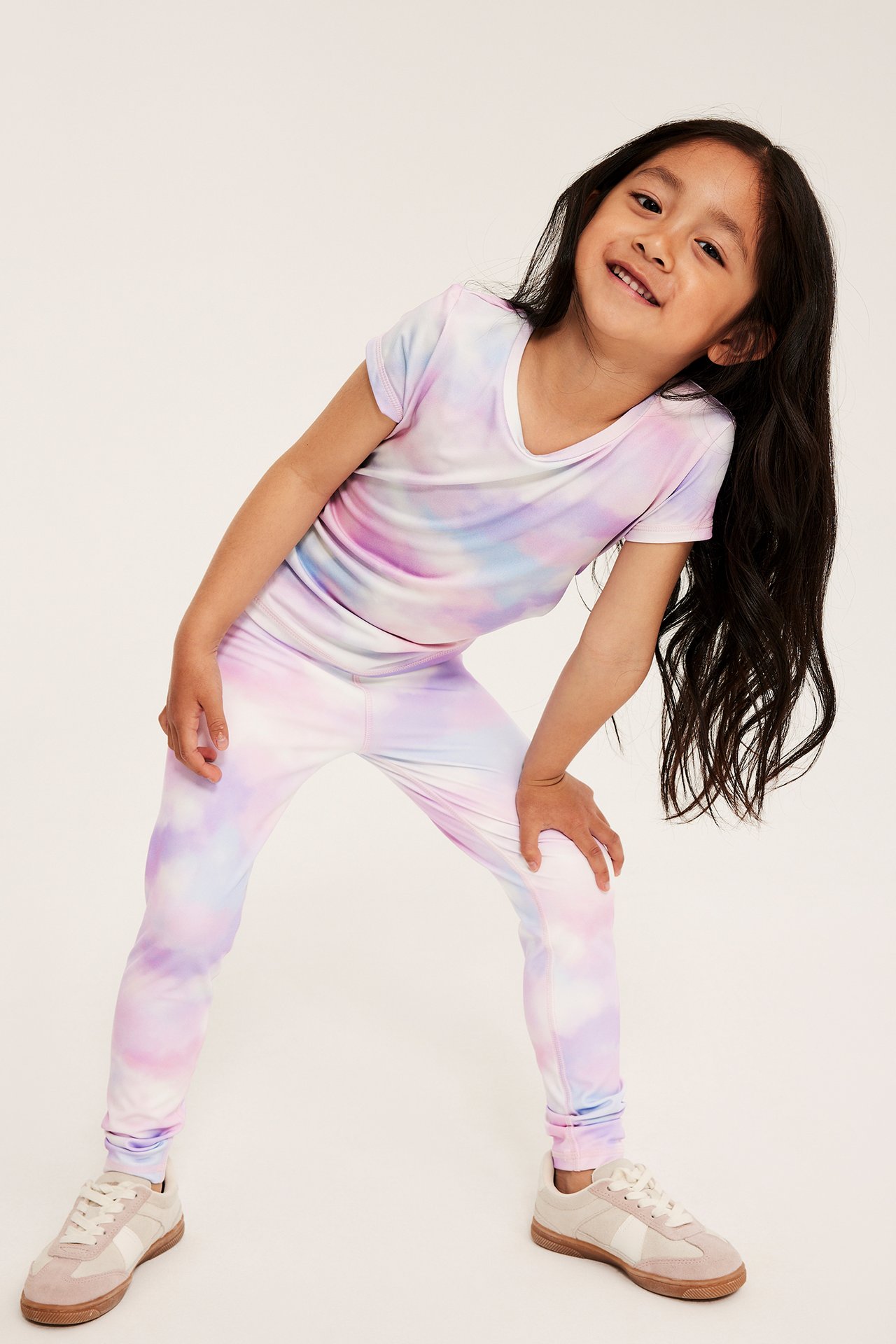 Edestä: Lapsella on luonnonvalkoiset tie dye -kuvioiset treenileggingsit, yhteensopiva V-paita ja beiget tennarit.