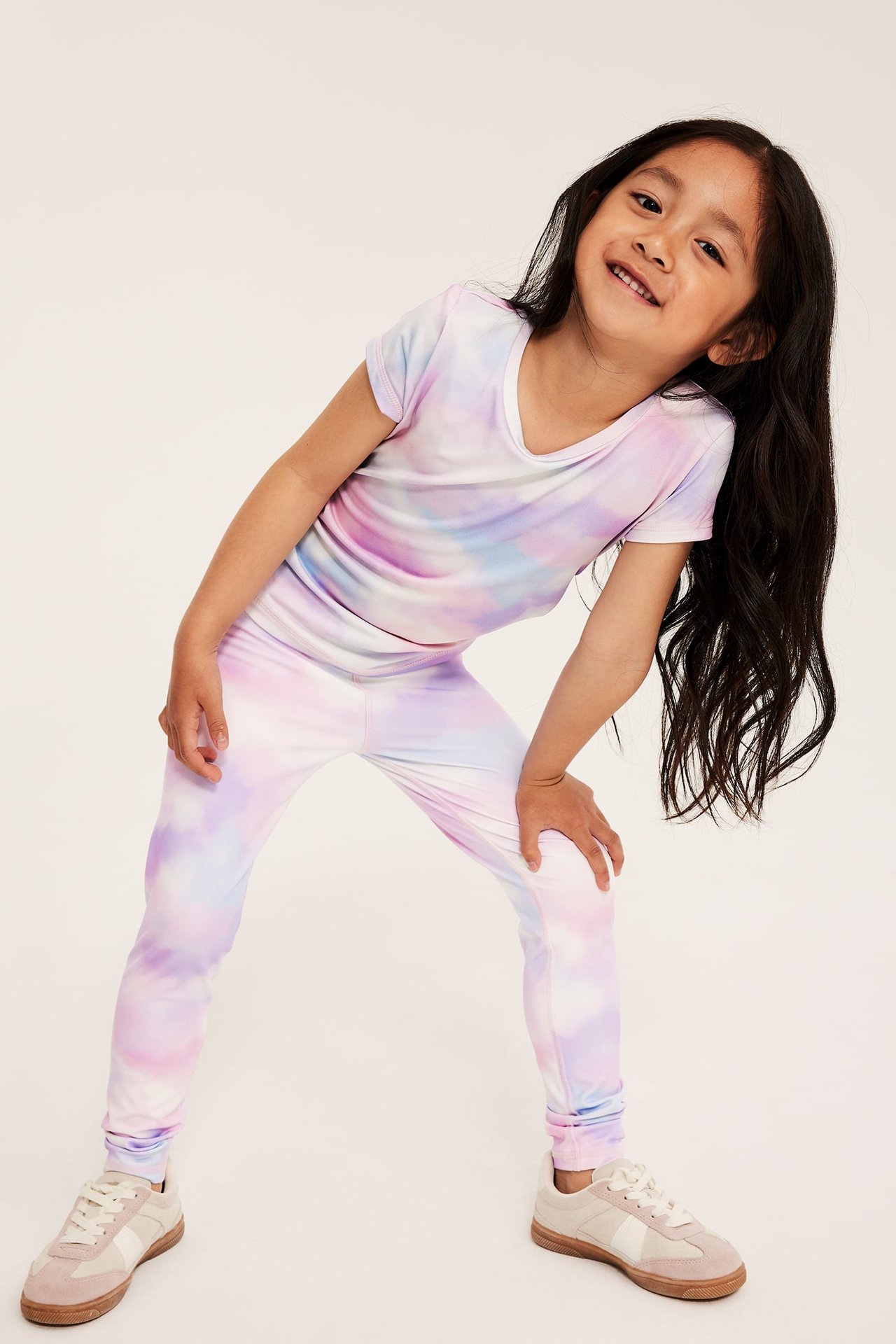 Widok z przodu: Dziecko nosi białawe legginsy treningowe tie-dye, pasujący T-shirt z dekoltem V i beżowe sneakersy.
