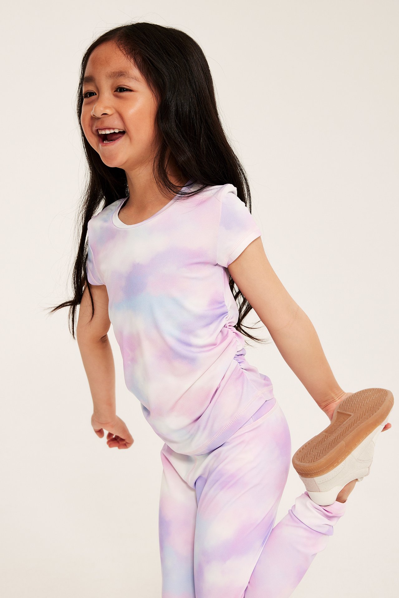 Edestäpäin hymyilevä lapsi tie-dye-kuvioisessa urheilutopissa rypytyksillä, yhteensopivissa leggingseissä ja valkoisissa tennareissa.