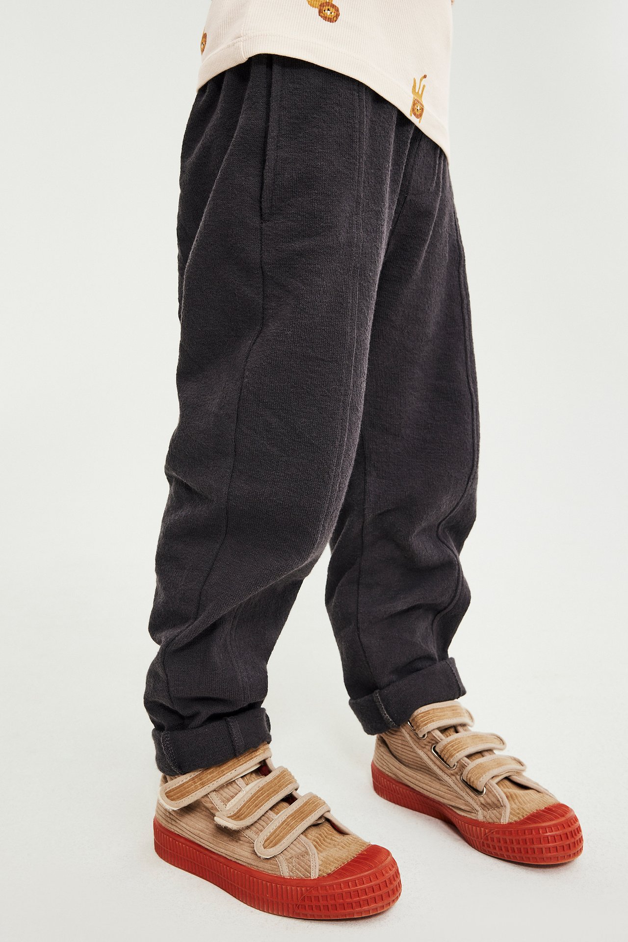 Närbild framifrån på barn i mörkgrå avslappnade sweatpants och beiga manchester sneakers med röda sulor.