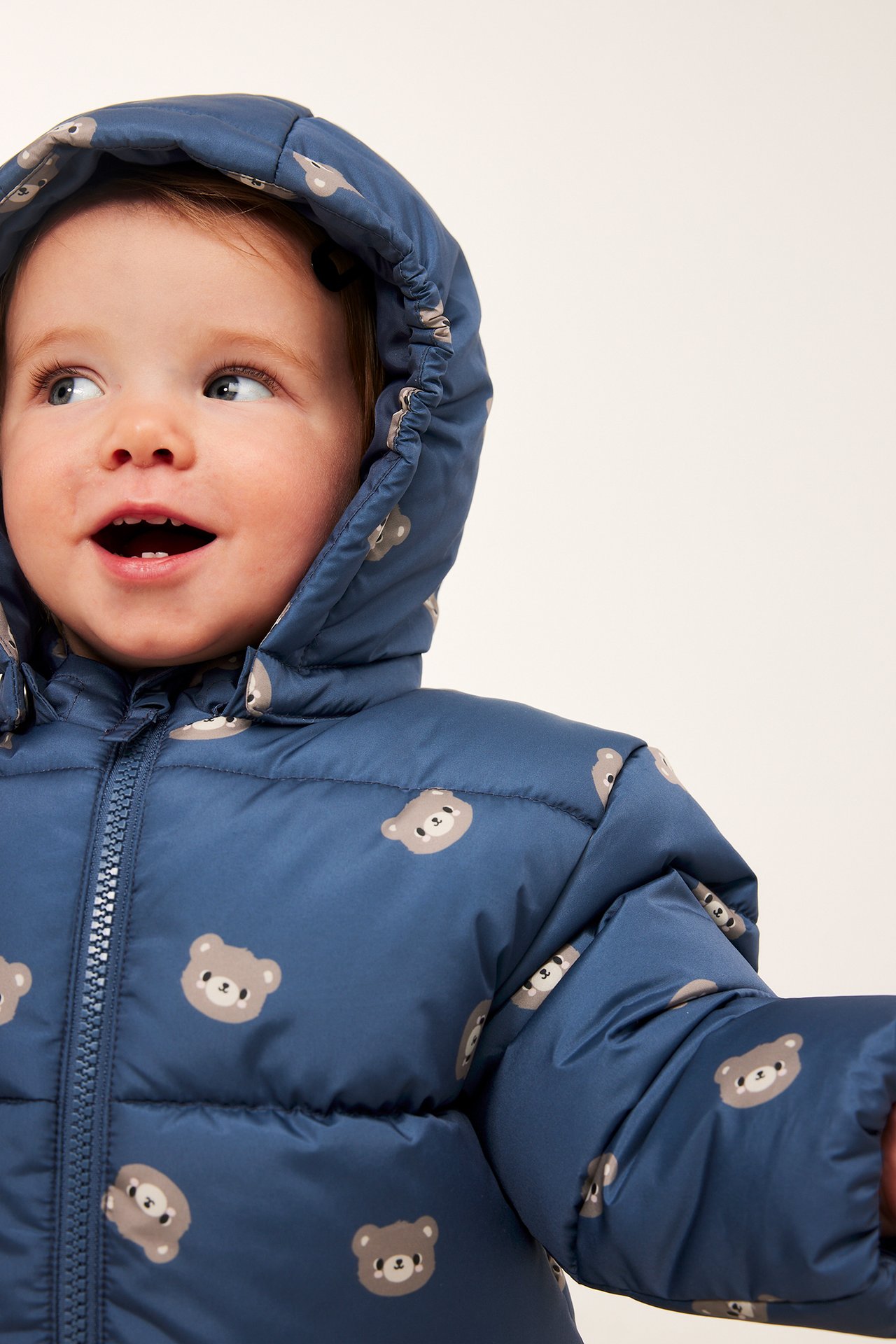 Närbild på en baby i en blå huvförsedd pufferjacka med brunt björnmönster och dragkedja.