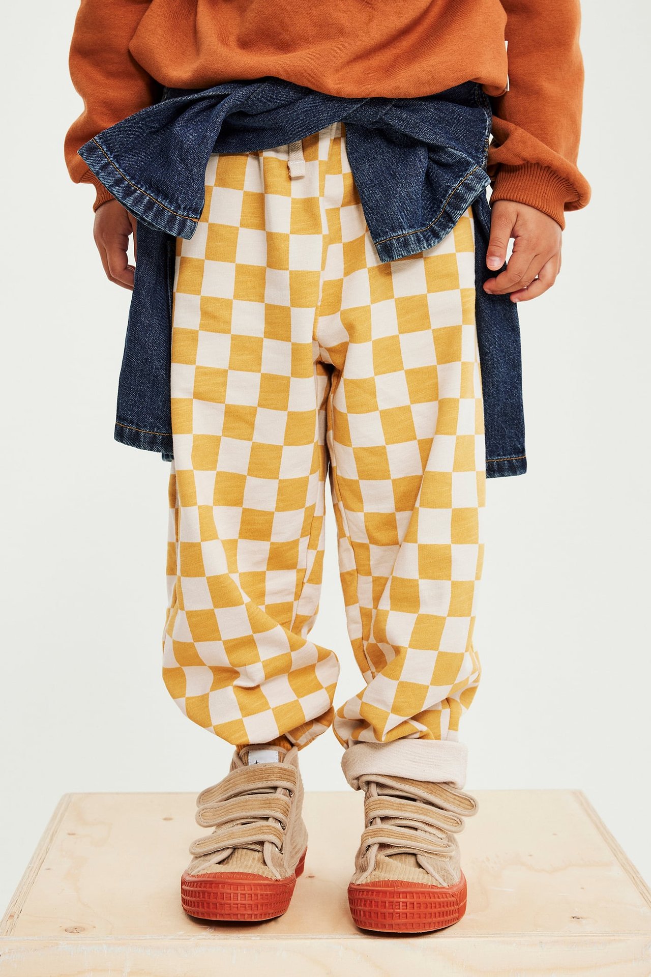 Frontbilde av barn i gule og hvitrutete joggebukser, oransje genser, dongerijakke og beige joggesko.