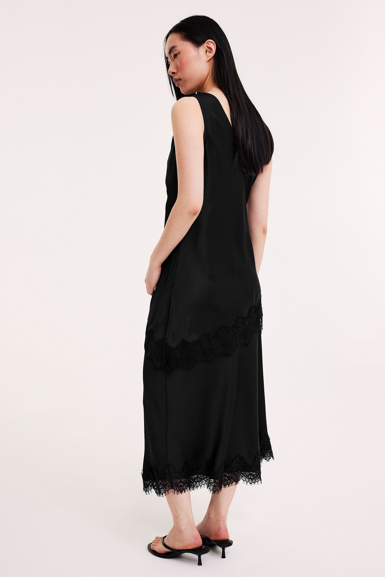 Sida vy av en kvinna i svart långt satinlinne med spetsdetaljer och svarta klack sandaler.