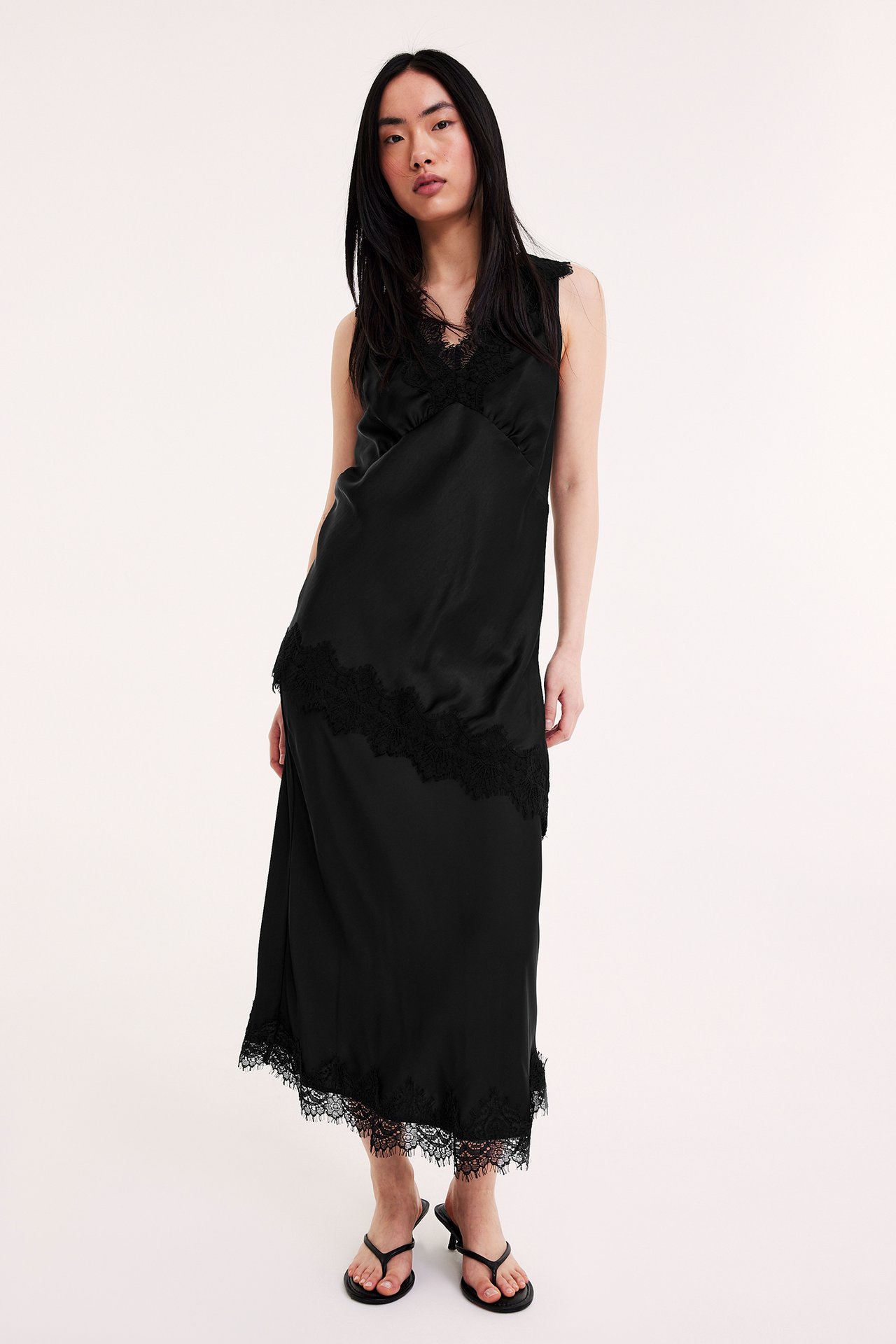 Frontvy av en kvinna i en svart lång slipklänning i satin med spetsdetaljer och svarta klackflipflops.