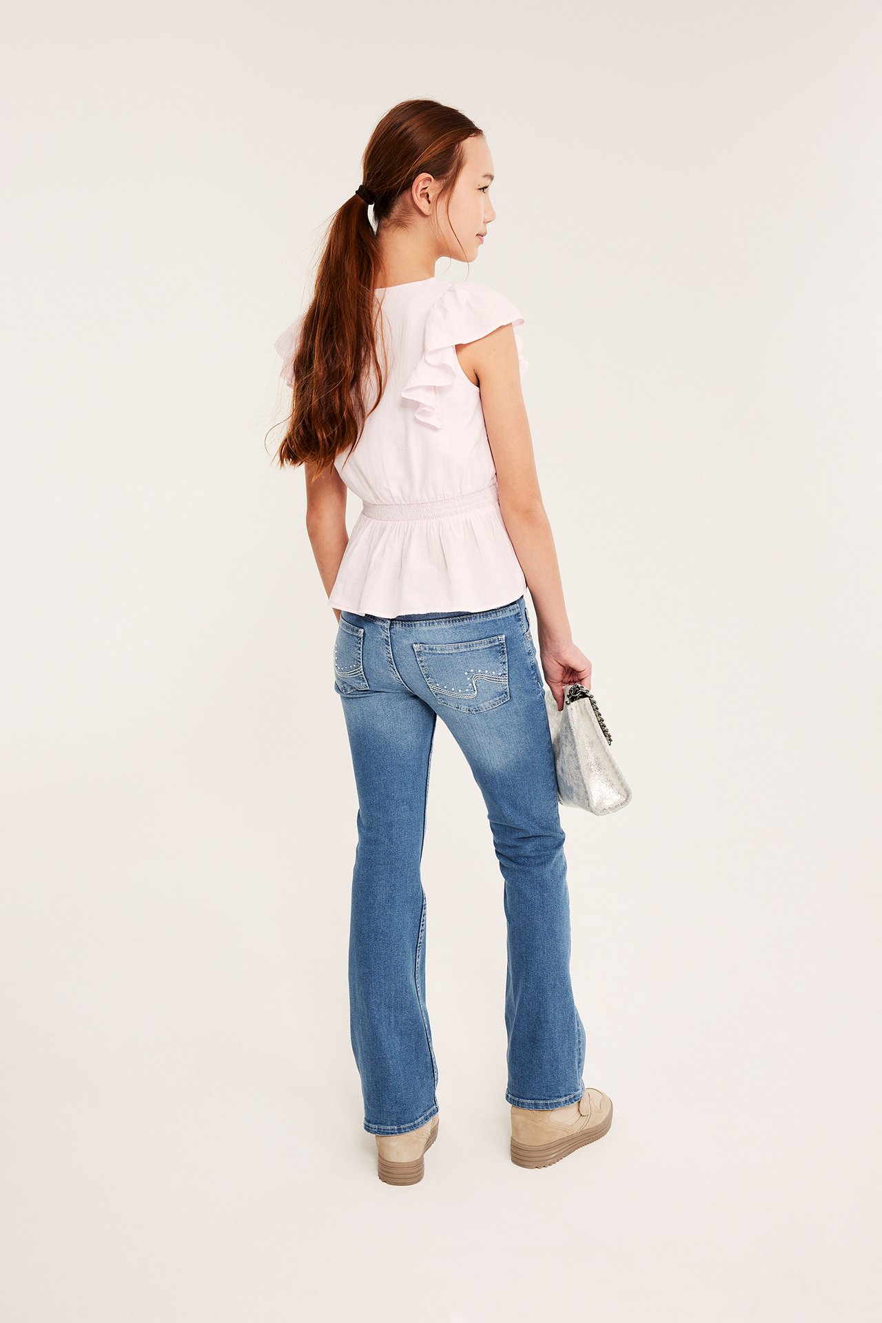 Widok z tyłu: Dziecko w jasnoniebieskich zdobionych jeansach bootcut, różowym topie, beżowych butach, trzyma srebrną torbę.