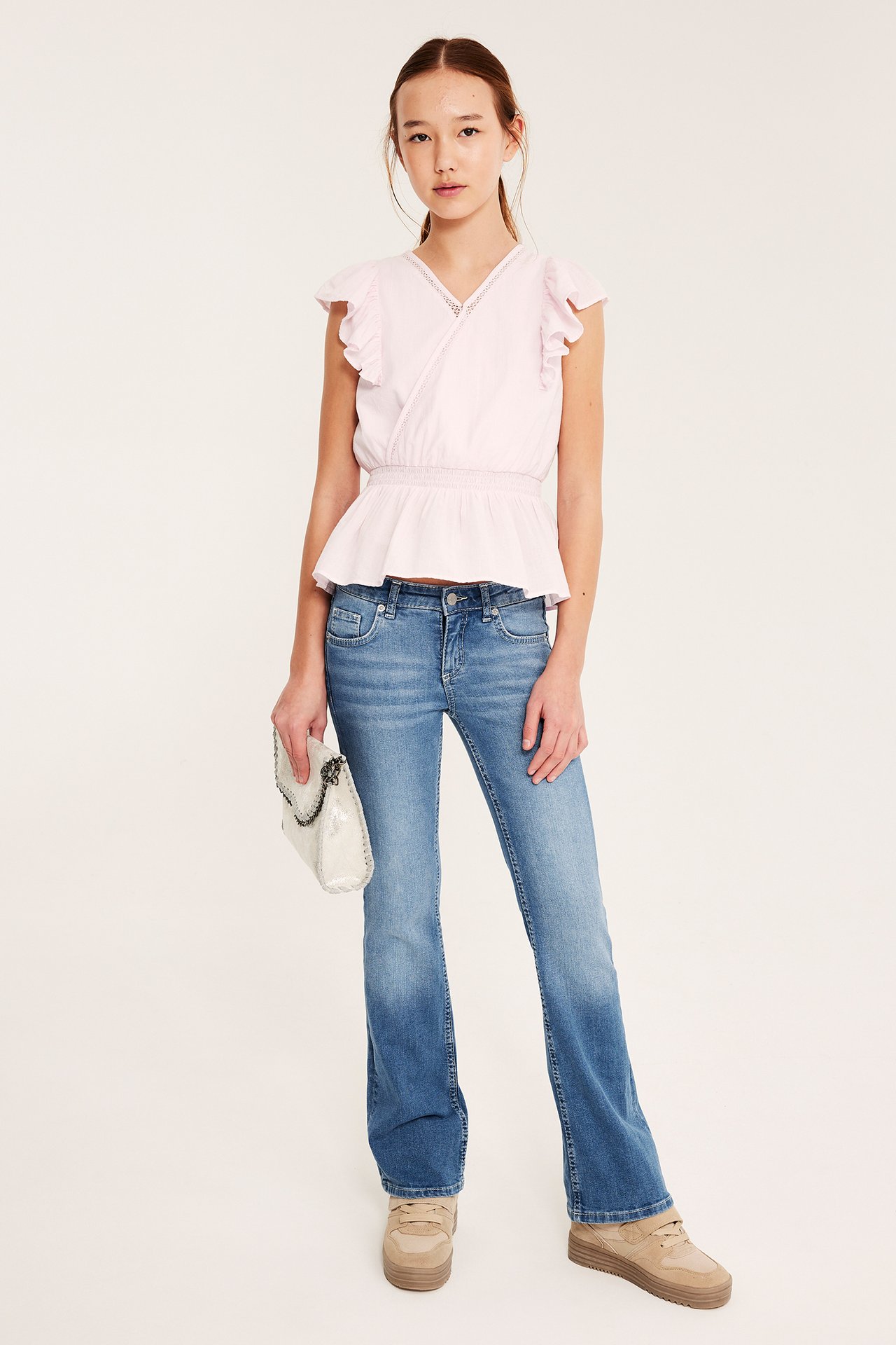 Edestä: lapsi vaaleansinisissä bootcut-farkuissa, vaaleanpunaisessa topissa, beigeissä kengissä, valkoista laukkua pitäen.