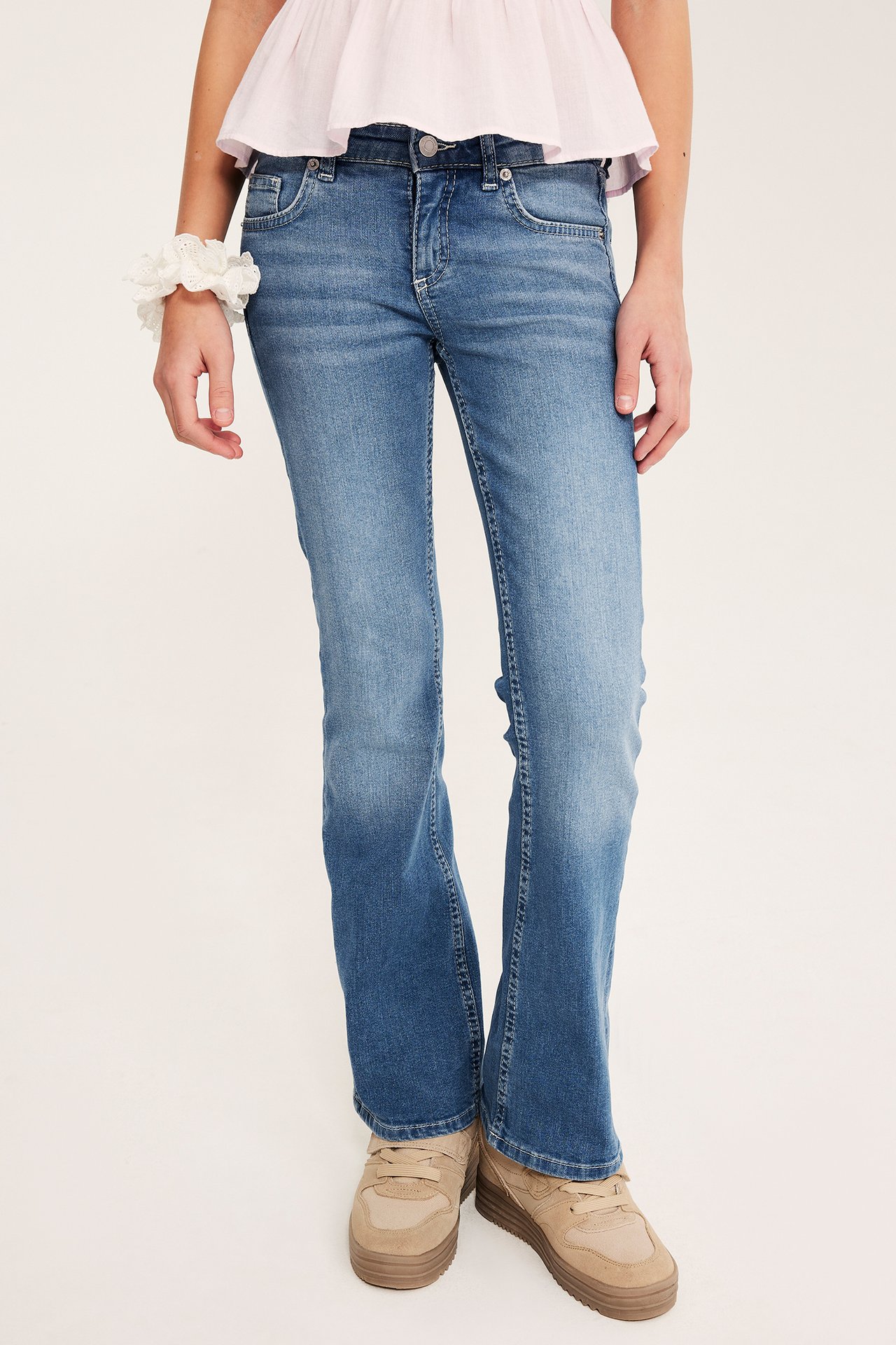 Frontvy: Barn bär ljusblå bootcut-jeans med låg midja, rosa topp, vit scrunchie och beiga sneakers.