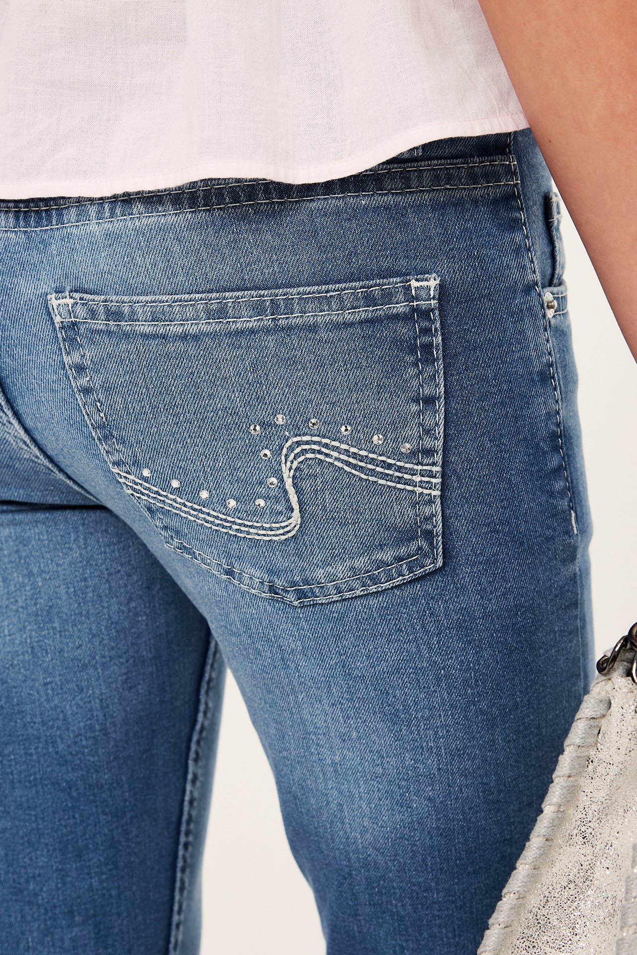 Närbild bakifrån: Kvinna i ljusblå bootcutjeans med dekorerade bakfickor, rosa topp, håller i en metallicväska.