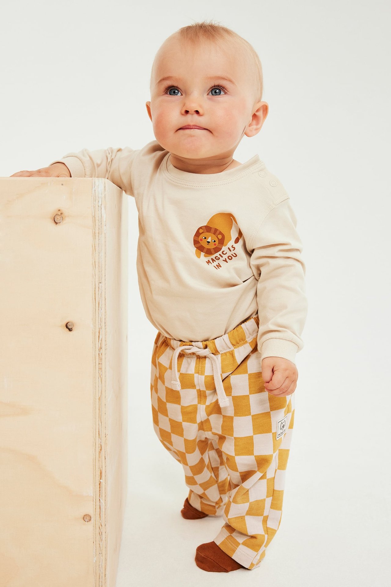 Frontbilde av baby i beige langermet topp med løvegrafikk og gulrutete bukser.