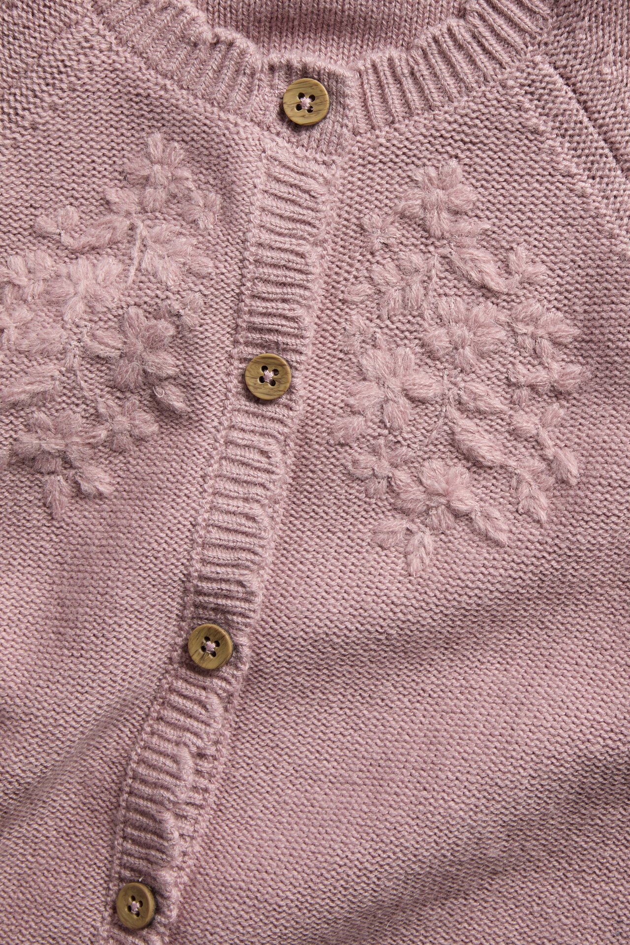 Närbild på en dammlila stickad kofta med blommigt broderi och träknappar.