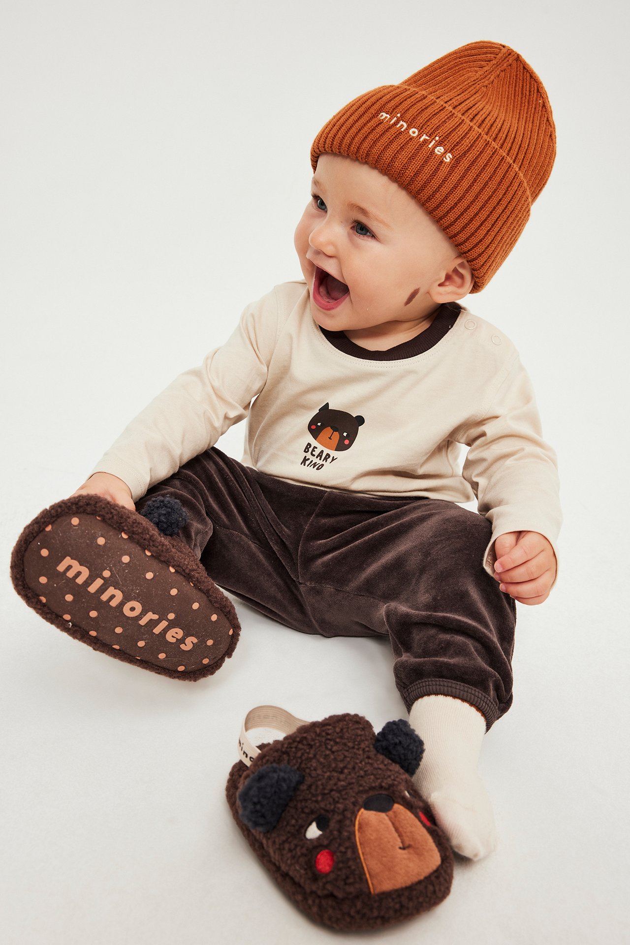 Edestä kuvattu vauva yllään ruosteenruskea 'minories' pipo, beige 'BEARY KIND' t-paita, ruskeat samettihousut ja ruskeat nallelapikkaat.